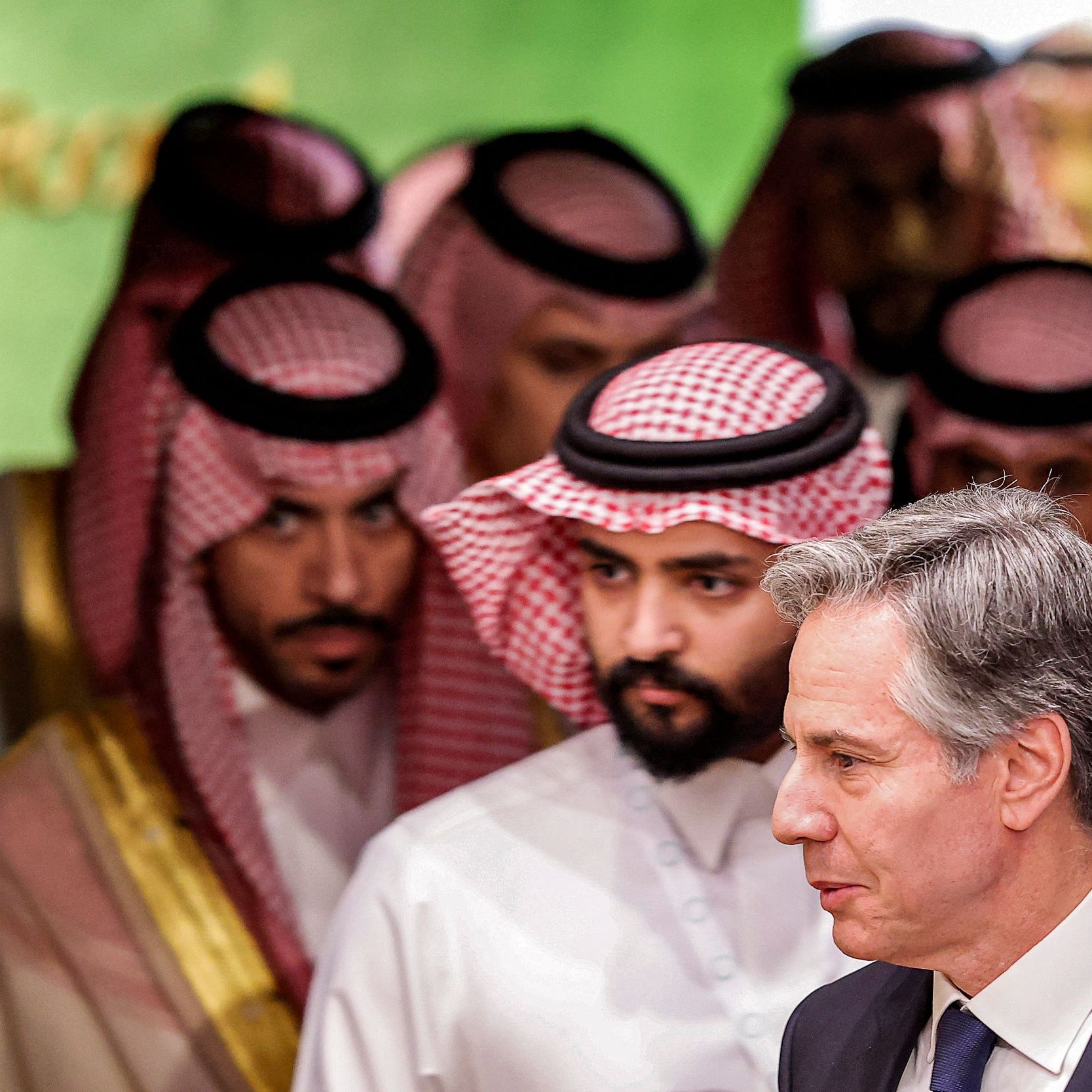 در این تصویر، تعدادی مرد که به نظر می‌رسد لباس‌های سنتی عربی پوشیده‌اند، در پس‌زمینه‌ای روشن و زنده قرار دارند. در جلو، مردی با کت و کراوات به سمت راست در حال حرکت است. به نظر می‌رسد این تصویر مربوط به یک نشست یا مراسم رسمی باشد. فضای کلی تصویر حس تعامل و گفت‌وگو را منتقل می‌کند.