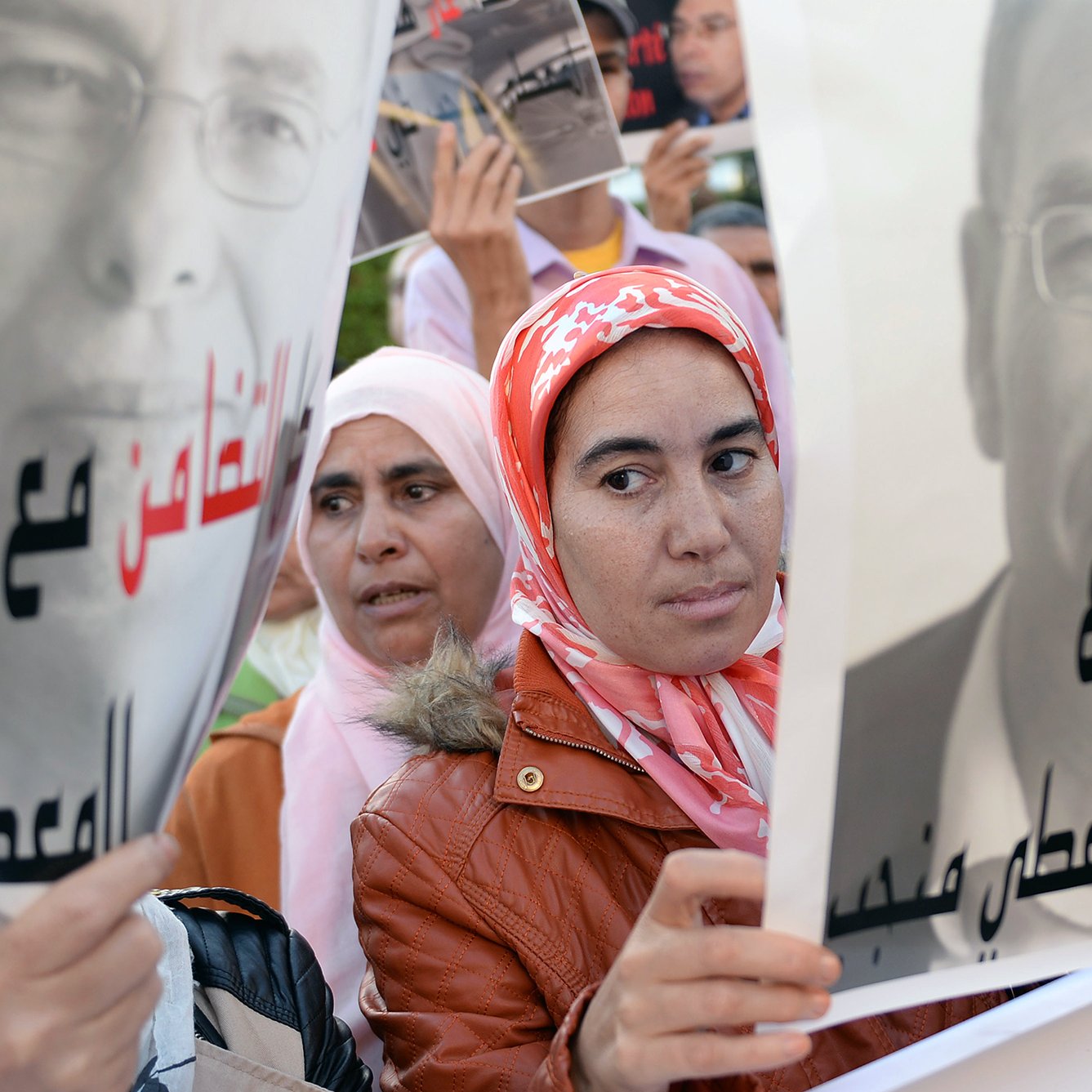 L'image présente des manifestants tenant des pancartes. Au premier plan, une femme avec un foulard rose regarde l'objectif, tandis qu'une autre femme, en arrière-plan, semble également attentive. Les pancartes affichent des portraits en noir et blanc d'un homme, accompagnés de slogans en arabe. L'ambiance suggère un rassemblement ou une manifestation, probablement pour défendre une cause ou exprimer une opinion.