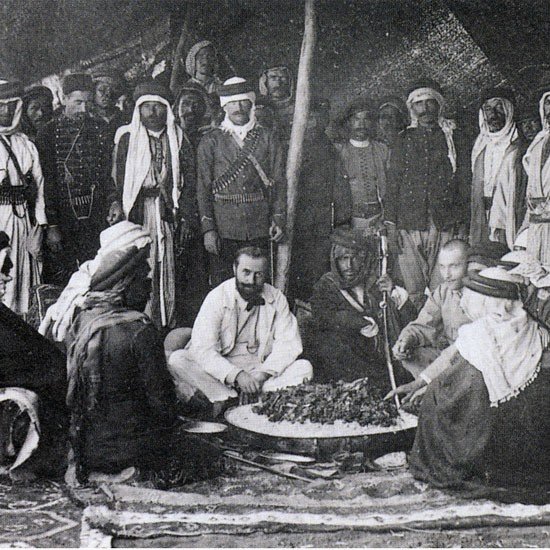 L'image montre un groupe de personnes, probablement issus de cultures arabes, rassemblées autour d'un grand plat de nourriture posé au sol. Certains sont assis tandis que d'autres se tiennent debout en arrière-plan. Les participants portent des vêtements traditionnels, tels que des robes et des turbans, ce qui suggère un contexte historique. L'ambiance semble conviviale, et l'événement pourrait être un repas ou une célébration. Des éléments du décor indiquent que la scène se déroule en extérieur, possiblement sous une tente.