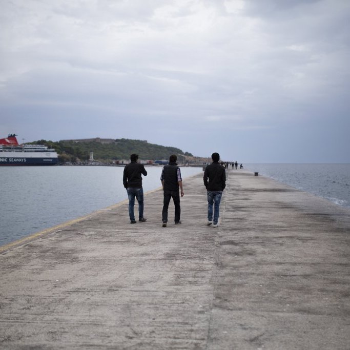 L'image montre trois personnes marchant sur un quai. Le ciel est nuageux, et l'atmosphère est calme. À l'arrière-plan, on peut voir un ferry amarré et une petite île. Le quai est en béton, et l'eau semble calme, reflétant les nuages. Les silhouettes des gens ajoutent une touche de vie à cette scène paisible.