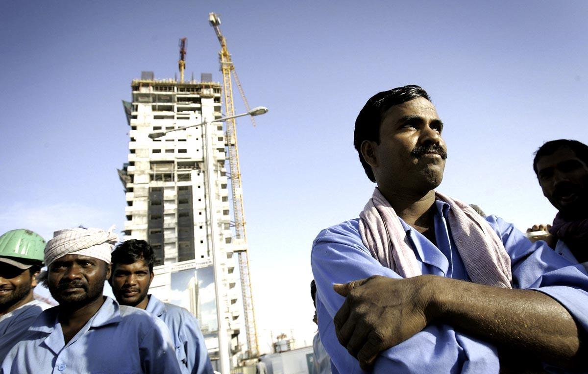 L'image montre un groupe d'ouvriers sur un chantier de construction. Au premier plan, un homme est au centre de l'image, les bras croisés et affichant une expression confiante. À l'arrière-plan, on voit un bâtiment en construction, avec des grues qui travaillent. Les ouvriers portent des vêtements de travail, et certains d'entre eux portent des casques de sécurité. Le ciel est dégagé, suggérant une journée ensoleillée. L'ensemble de la scène évoque l'atmosphère d'un site de construction animé avec des travailleurs engagés dans leur tâches.