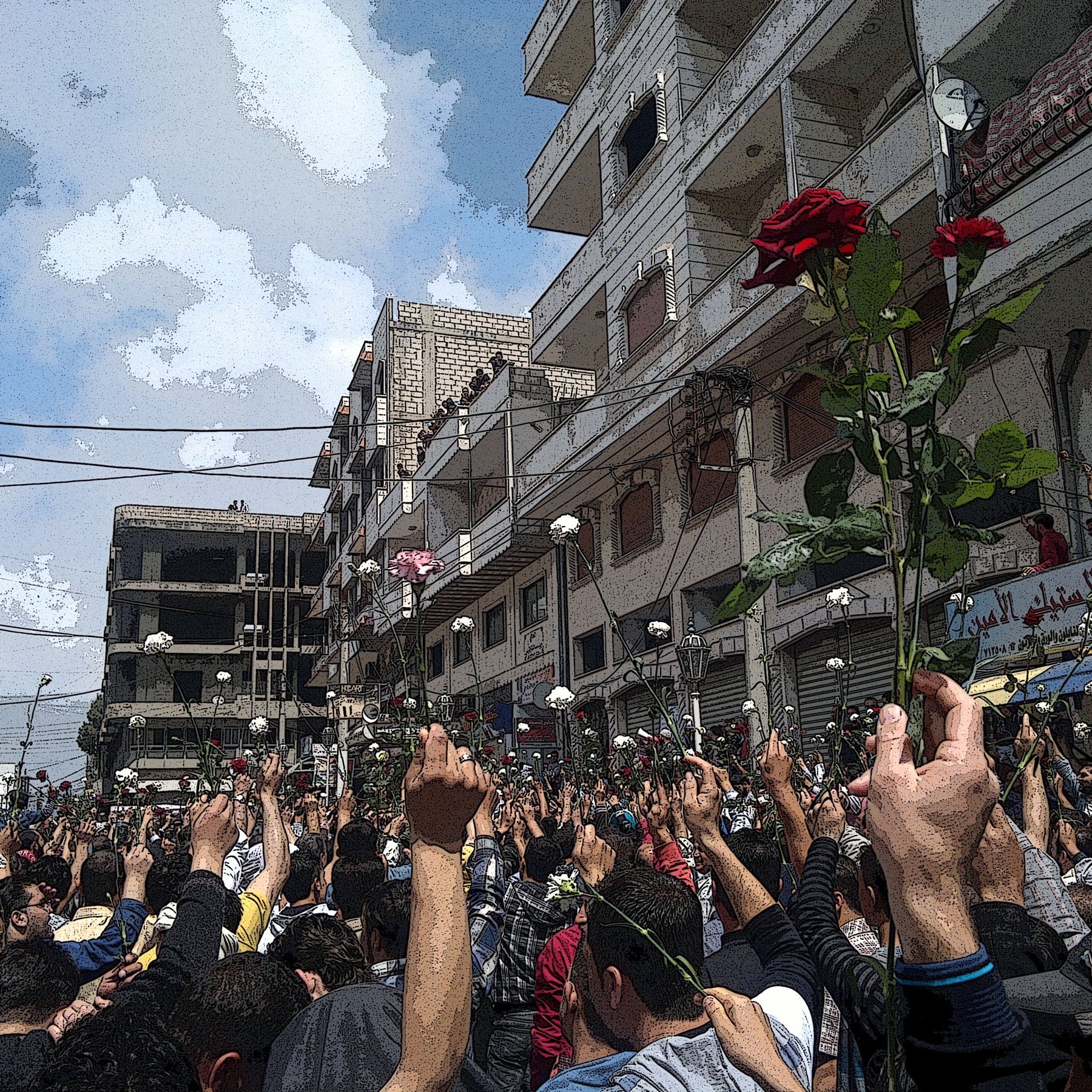 L'image montre une grande foule au milieu d'une rue, probablement lors d'une manifestation. Les gens semblent levés les bras, tenant des roses, ce qui suggère une ambiance pacifique ou festive. En arrière-plan, on aperçoit des bâtiments et un ciel partiellement nuageux. L'atmosphère semble être chargée d'émotions et d'unité entre les participants.