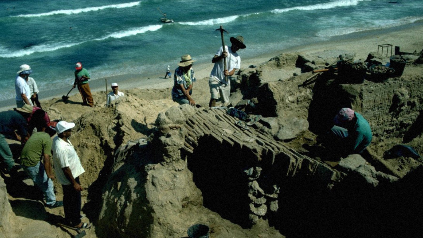Des archéologues creusent sur une plage, entourés de sable et de vagues.