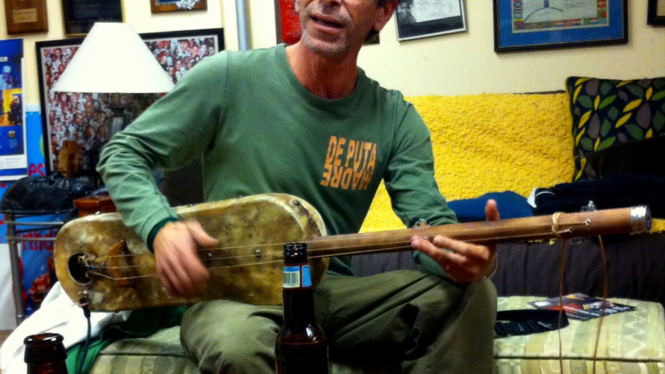 L'image montre un homme assis dans un intérieur chaleureux, probablement un salon. Il joue d'un instrument de musique, semblable à une guitare, tout en chantant. Il porte un t-shirt avec un message audacieux et des pantalons clairs. À côté de lui, sur une table basse, on aperçoit quelques bouteilles de bière. Les murs autour sont décorés de plaques et de diplômes, et un canapé coloré est visible en arrière-plan. L'ambiance semble conviviale et détendue.