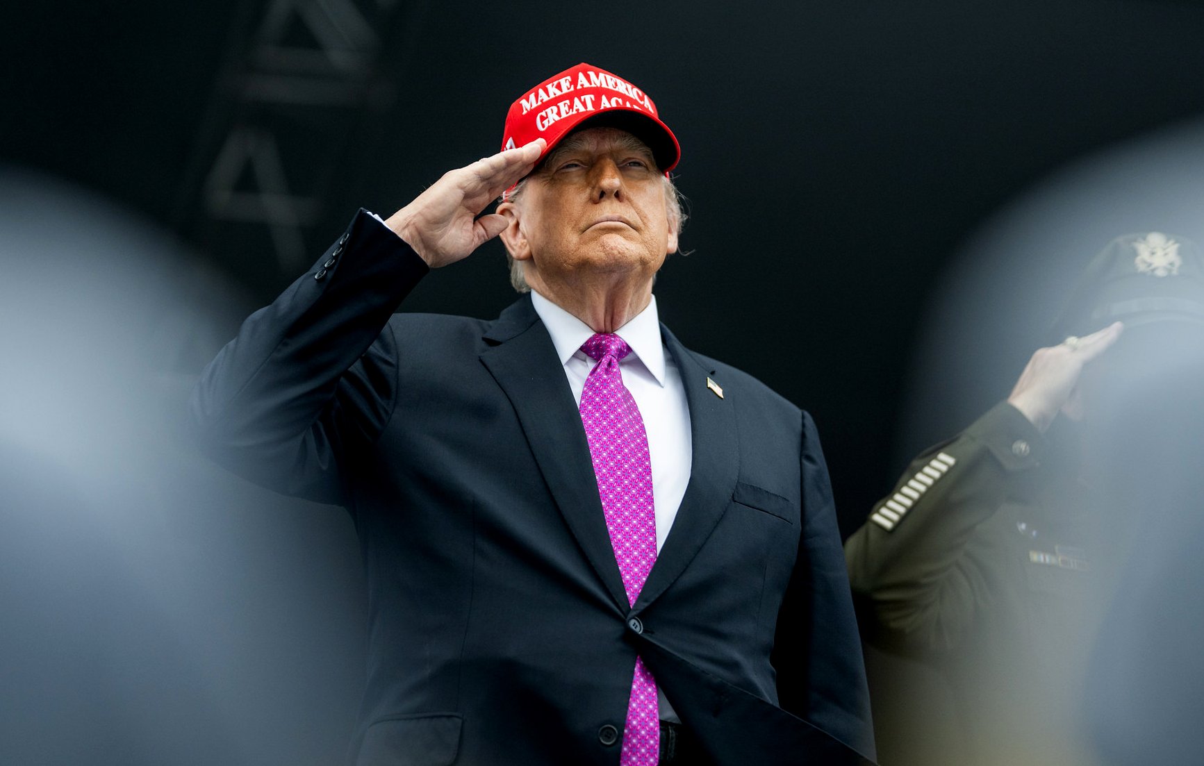 Homme en costume noir, portant une casquette rouge, effectuant un salut.