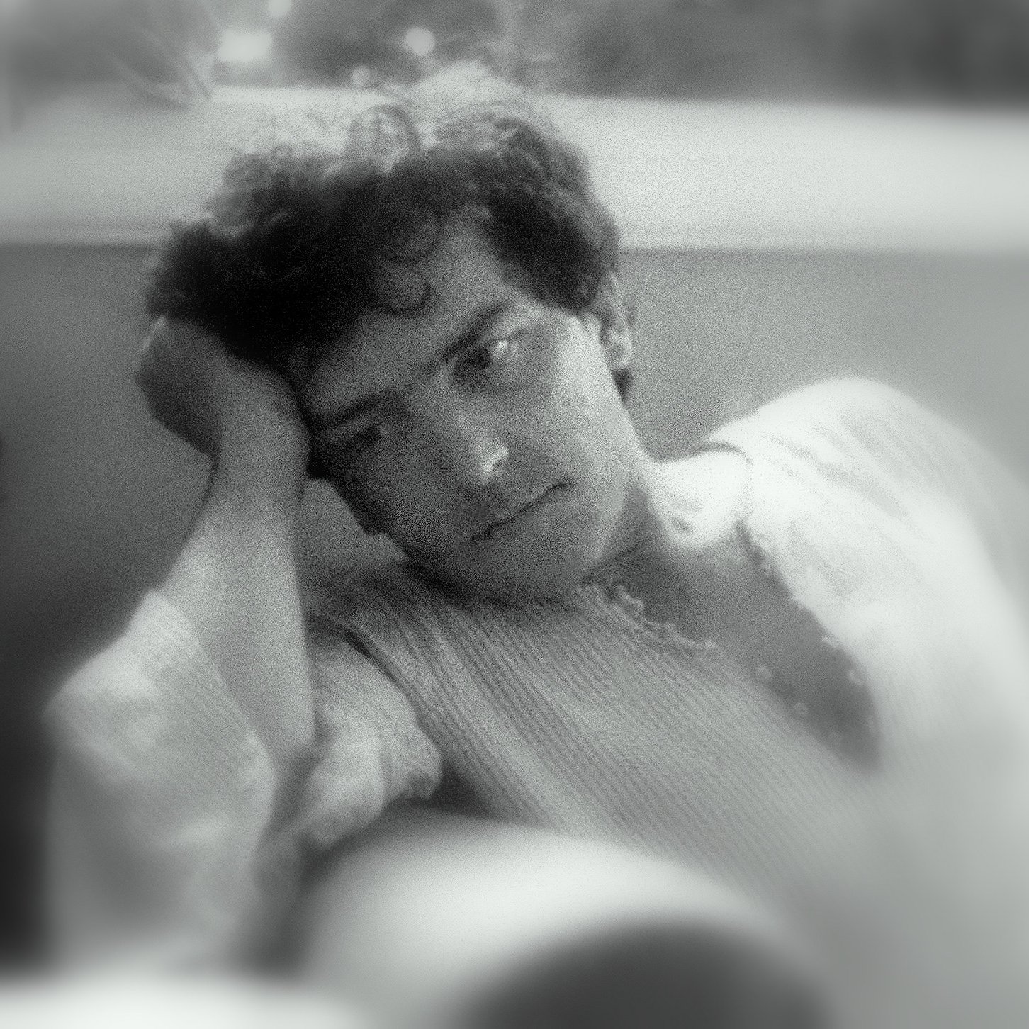 L'image montre un homme pensif, assis dans un endroit confortable. Il a les cheveux bouclés et un air réfléchissant, avec une main soutenant sa tête. Son expression semble mélancolique ou pensive. L'éclairage doux donne une ambiance nostalgique et calme à la scène. L'arrière-plan flou contribue à centrer l'attention sur son visage.