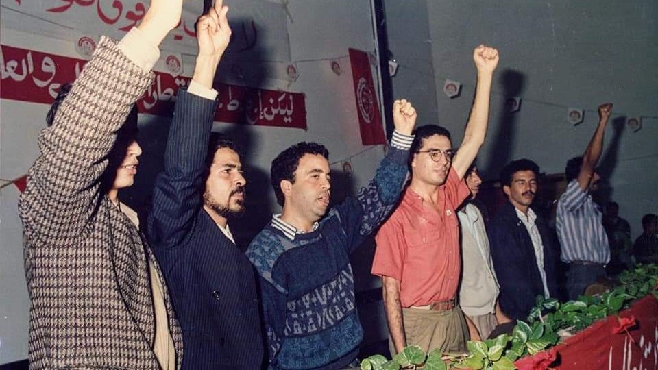 تظهر الصورة مجموعة من الأشخاص واقفين خلف منصة، يرفعون أيديهم في إشارة للتعبير عن الاحتجاج أو الدعم. الخلفية تحتوي على لافتات مكتوبة، مما يشير إلى أن الحدث قد يكون سياسيًا أو اجتماعيًا. الجو العام يبدو حماسيًا، حيث يعكس تفاعل الجمهور مع ما يحدث أمامهم. الأشخاص يرتدون ملابس تتنوع في الألوان والأساليب، مما يعكس حقبة زمنية معينة.