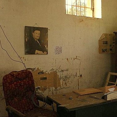 L'image montre une pièce semblant abandonnée ou négligée. Sur le mur, il y a une photo encadrée d'un homme. Le décor comprend une chaise rouge en velours, une table en bois avec quelques objets dessus, et une vitrine en bois remplie de divers éléments. Les murs sont marqués par l'usure et présentent des traces de détérioration. La lumière pénètre à travers une fenêtre, illuminant partiellement l'espace. L'atmosphère générale est nostalgique et témoigne d'un passé révolu.