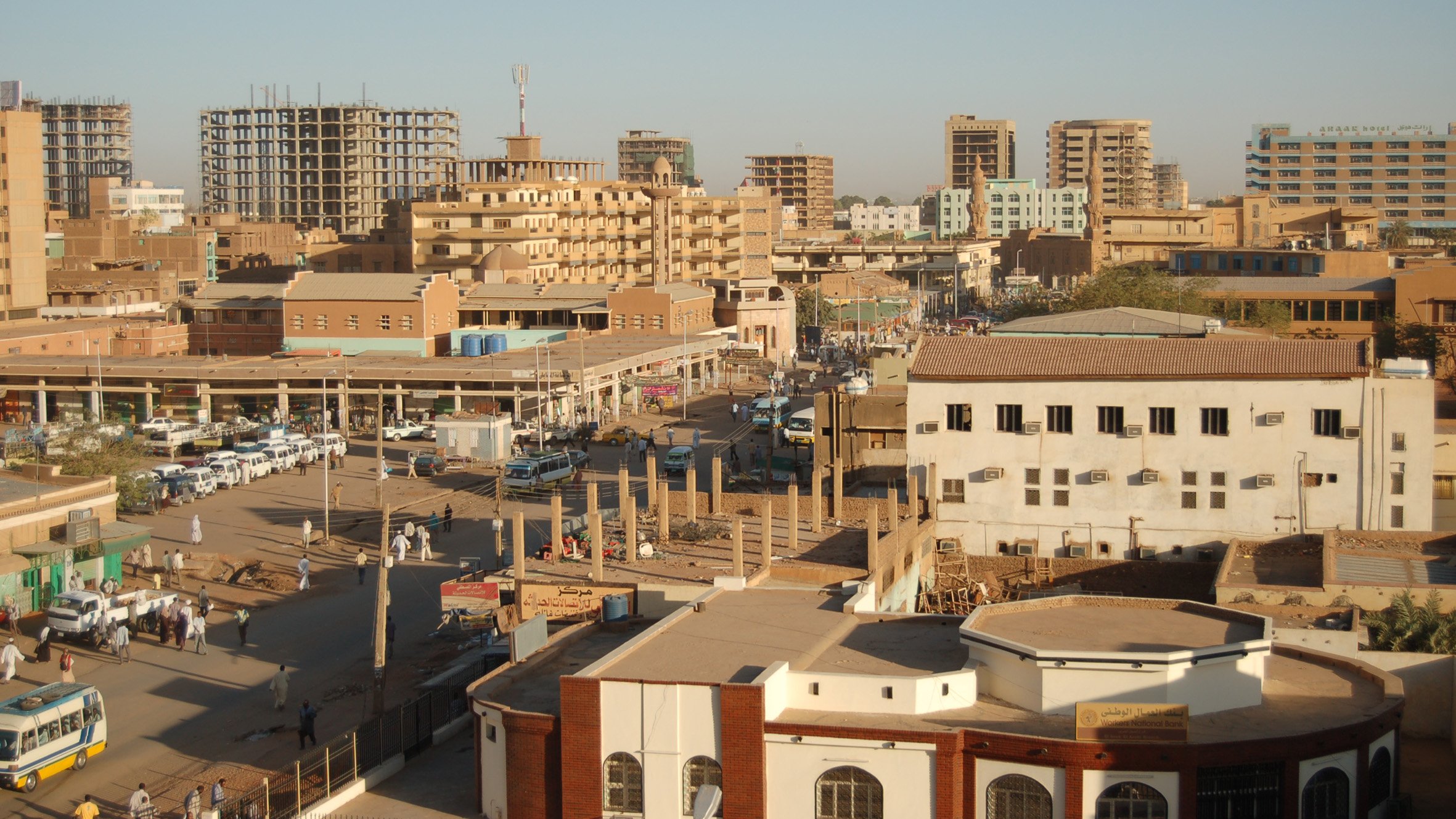 L'image montre une vue panoramique d'une ville, probablement une ville nord-africaine ou du Moyen-Orient. On peut voir plusieurs bâtiments, certains modernes et d'autres plus anciens, ainsi que des structures en construction. La rue est animée avec des voitures et des gens qui se déplacent. L'architecture varie, avec un mélange de styles. Le ciel est dégagé, indiquant probablement une belle journée ensoleillée. Les bâtiments sont de différentes hauteurs et couleurs, donnant à la scène un aspect dynamique et vivant.