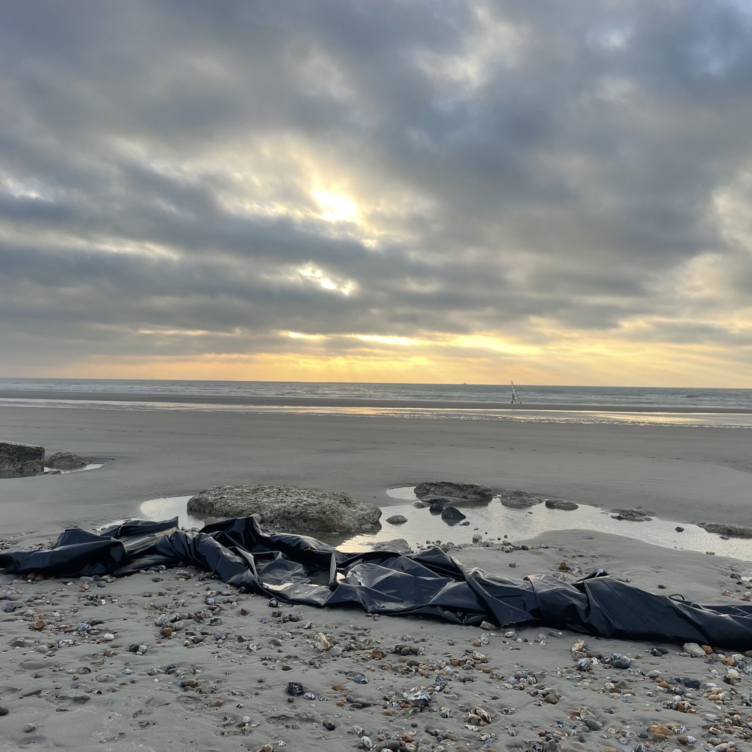 L'image montre une plage désertique au crépuscule, avec un ciel nuageux où quelques rayons de soleil percent à travers les nuages. Sur le sable, on aperçoit un long objet noir qui semble être un morceau de toile ou de bâche, posé près de quelques rochers. À l'horizon, la mer et quelques petites vagues reflètent la lumière tamisée du ciel. L'atmosphère est à la fois calme et mystérieuse.