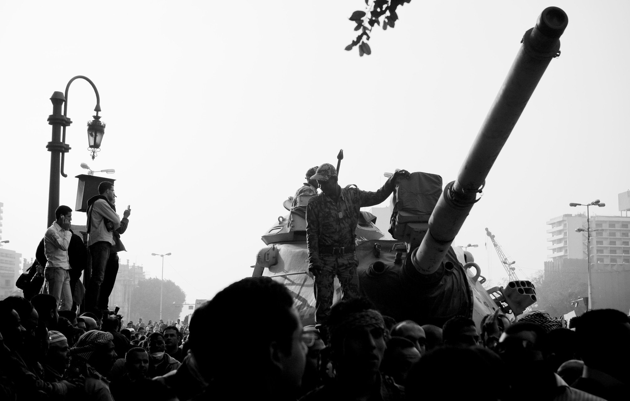 L'image montre une scène de manifestation ou de rassemblement avec une atmosphère intense. Au centre, on peut voir un soldat debout sur un char, levant le bras en signe de défi ou de puissance. Le char est entouré d'une foule dense de personnes, certaines levant les bras ou prenant des photos. L'arrière-plan semble urbain, avec des bâtiments et un lampadaire. La photo est en noir et blanc, ce qui ajoute à la dramaturgie et à la tension de la scène.
