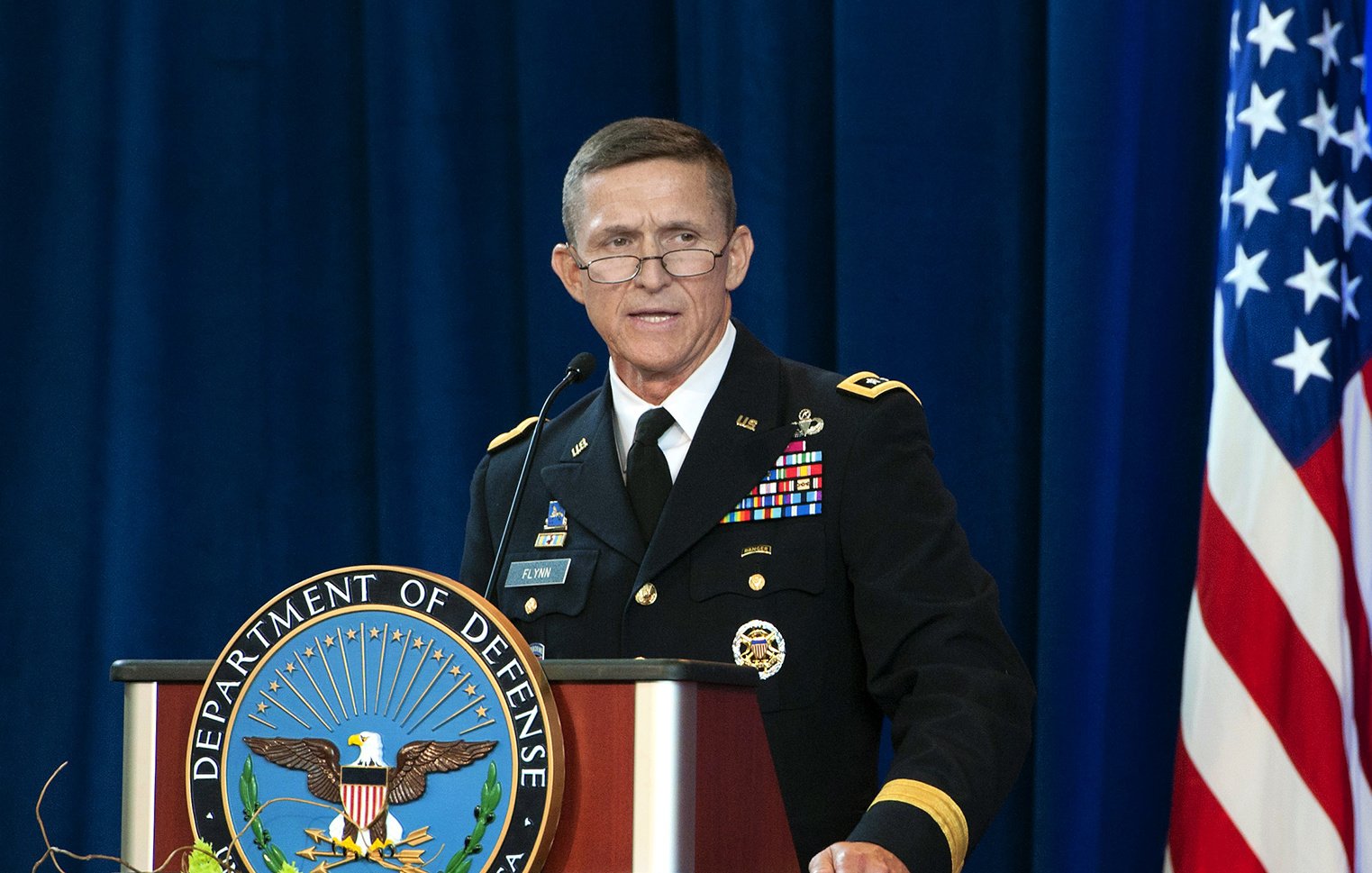 The image shows a man in a military uniform standing at a podium. The podium has the emblem of the Department of Defense prominently displayed. The man is speaking, possibly at a formal event or press conference. Behind him, there is a blue backdrop, and an American flag is visible to the side. The setting appears to be official and ceremonial.