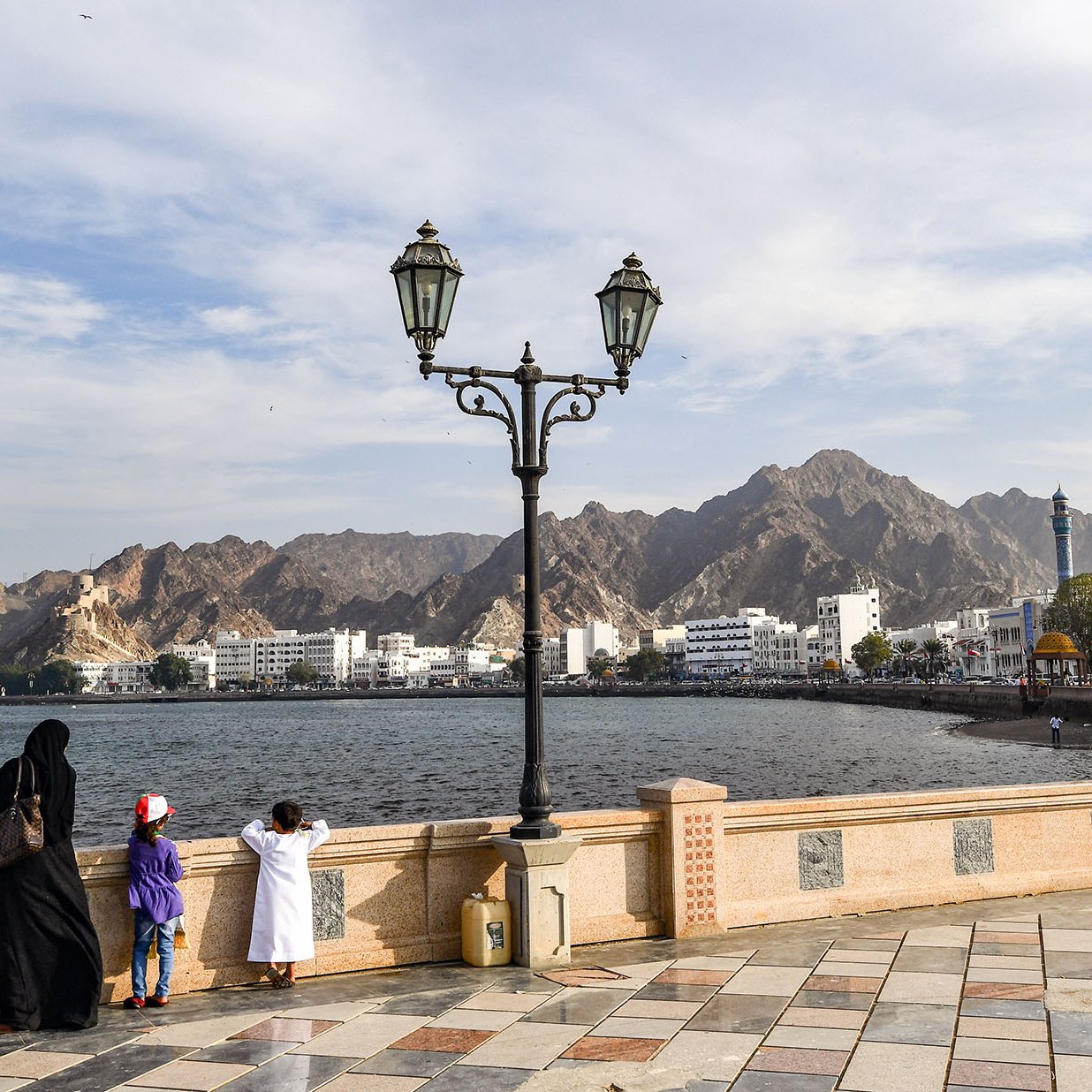 L'image montre une scène de bord de mer, probablement à Muscat, Oman. Dans le premier plan, on peut voir une famille vêtue de vêtements traditionnels. Un homme et une femme se tiennent près d'un lampadaire, regardant vers l'horizon. À côté d'eux, deux enfants, dont un porte une casquette rouge. En arrière-plan, on aperçoit des montagnes majestueuses ainsi que des bâtiments blancs typiques de la région. Le ciel est partiellement nuageux, créant une ambiance paisible et contemplative.