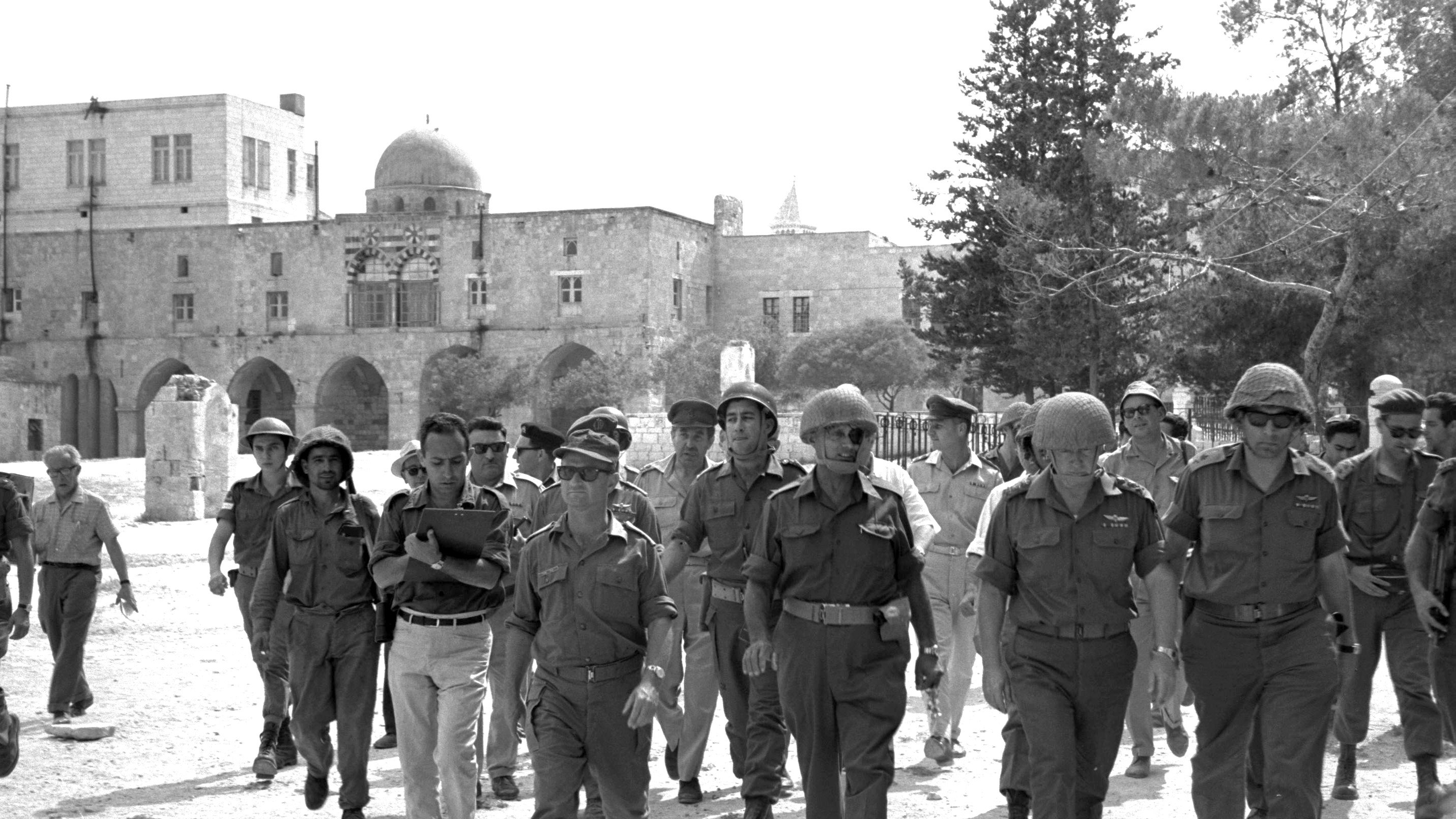 در این تصویر، گروهی از نظامیان و مأموران امنیتی در حال راه رفتن به سمت دوربین هستند. آن‌ها لباس‌های نظامی و کلاه‌های مخصوص به سر دارند. پس‌زمینه تصویر، شامل ساختمان‌های سنگی قدیمی و برخی درختان است. به نظر می‌رسد این تصویر در یک منطقه تاریخی یا باستانی گرفته شده باشد. نور خورشید به وضوح بر روی صحنه می‌تابد و فضایی گرم به تصویر می‌بخشد.