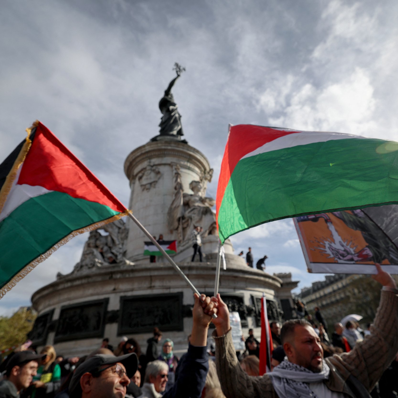 تظهر الصورة مجموعة من الأشخاص وهم يحملون أعلام فلسطينية في تجمع جماهيري. الخلفية تُظهر نصبًا تذكاريًا عظيمًا، مع سماء ملبدة بالغيوم. يبدو أن الناس متحمسون ويشاركون في فعالية ذات طابع وطني أو سياسي. الأعلام تحمل ألوان العلم الفلسطيني، مما يشير إلى دعمهم للقضية الفلسطينية.