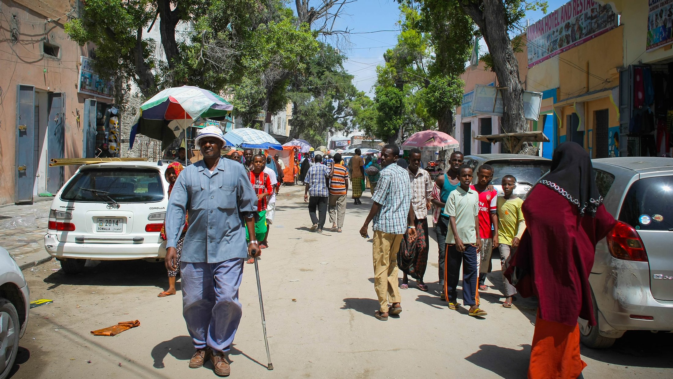 The image depicts a bustling street scene, likely in an urban area. There are several people walking along the road, some carrying umbrellas for shade. A few vehicles are parked along the sides, including cars that are mostly white. The atmosphere appears lively, with a variety of pedestrians, including a man using a walking cane. The buildings on either side are relatively simple, suggesting a community environment. Trees provide some greenery, enhancing the street's appearance. Overall, it captures a moment of daily life in a vibrant neighborhood.