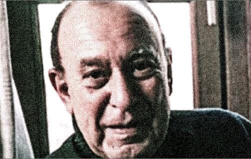 Je ne sais pas qui il est, mais l'image montre un homme âgé avec un sourire chaleureux. Il est éclairé par la lumière naturelle, et on peut voir des rideaux derrière lui. L'ambiance de l'image semble amicale et douce.