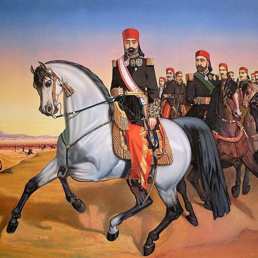 L'image représente un groupe de cavaliers en tenue traditionnelle, menés par un homme à cheval en avant-plan. Cet homme, qui semble être une figure d'autorité, est vêtu d'un uniforme orné et porte un chapeau distinctif. Il est monté sur un cheval gris, tandis qu'un groupe d'hommes sur des chevaux bruns et noirs le suit derrière lui. Le paysage est désertique, avec des montagnes en arrière-plan et un ciel aux teintes chaudes, suggérant le lever ou le coucher du soleil. Cette scène dégage une impression de mouvement et de puissance.