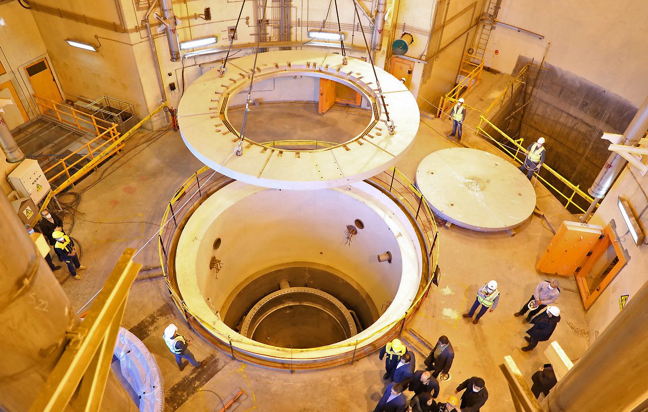 L'image montre l'intérieur d'une installation industrielle, probablement liée à la production d'énergie. On y voit un grand réacteur ou un réservoir cylindrique au centre, entouré de structures métalliques et de passerelles. Au-dessus, une grande plateforme circulaire est en train d'être soulevée, tandis qu'en bas, on aperçoit une zone vide. Plusieurs personnes en casques de sécurité se trouvent dans la pièce, probablement en train d'inspecter ou de superviser l'opération. L'éclairage est principalement artificiel, et les murs sont en béton. L'ensemble dégage une atmosphère technique et contrôlée.