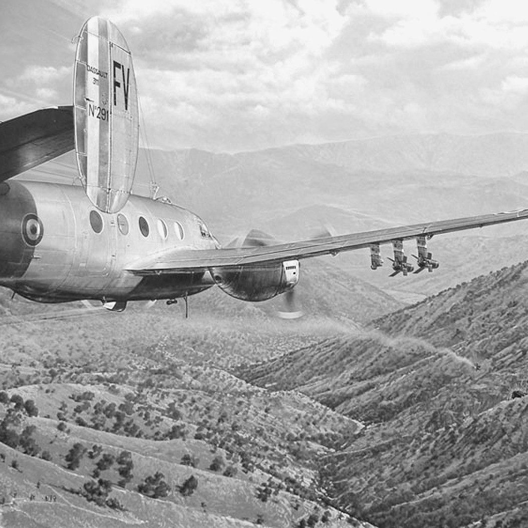 L'image montre un avion en vol, survolant un paysage montagneux. On peut voir des collines verdoyantes et des vallées en dessous, avec une atmosphère dramatique créée par les nuages. L'avion a un design classique, avec une queue distinctive et plusieurs réacteurs visibles. Ce type de scène évoque souvent des thèmes liés à l'aviation ou à la surveillance aérienne.