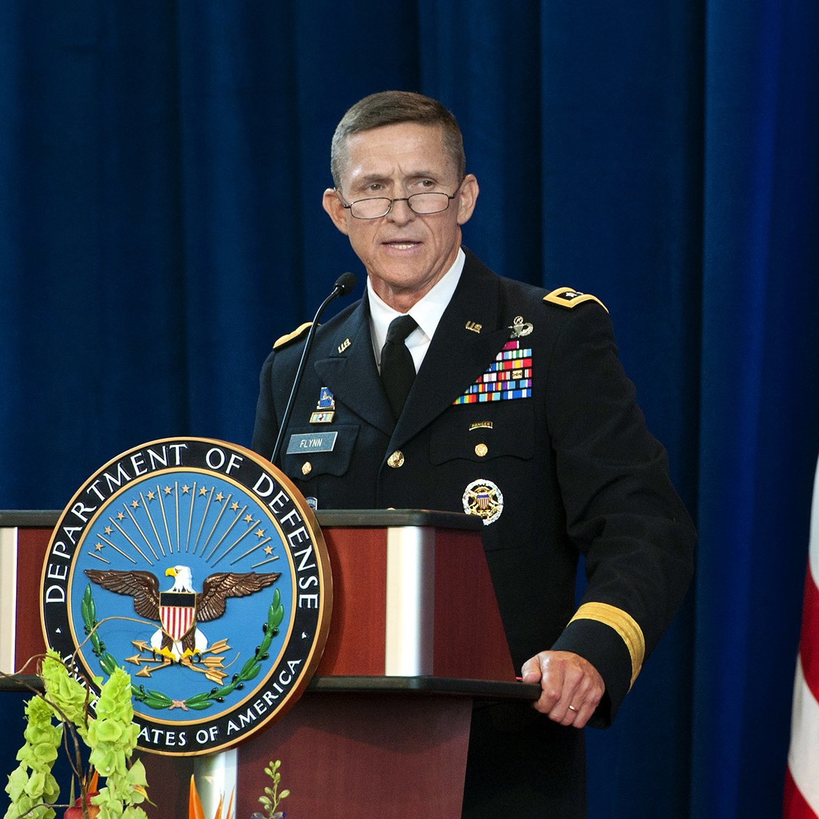 The image shows a man in a military uniform standing at a podium. The podium has the emblem of the Department of Defense prominently displayed. The man is speaking, possibly at a formal event or press conference. Behind him, there is a blue backdrop, and an American flag is visible to the side. The setting appears to be official and ceremonial.