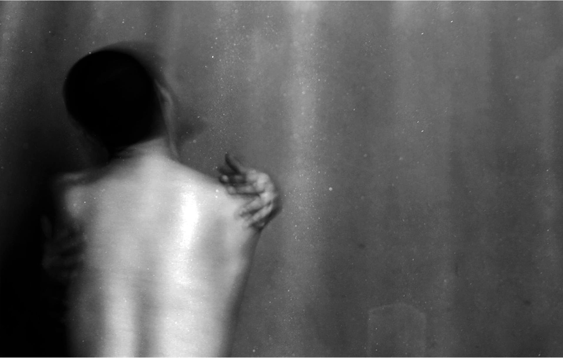 L'image montre une silhouette floue d'une personne de dos, avec un éclairage doux qui fait ressortir les contours de son corps. L'arrière-plan est sombre, créant une atmosphère intime et mystérieuse. La posture de la personne évoque une certaine vulnérabilité, tandis qu'une main semble s'accrocher à son épaule, suggérant une interaction ou un lien émotionnel. L'utilisation du noir et blanc renforce l'aspect dramatique et introspectif de la scène.