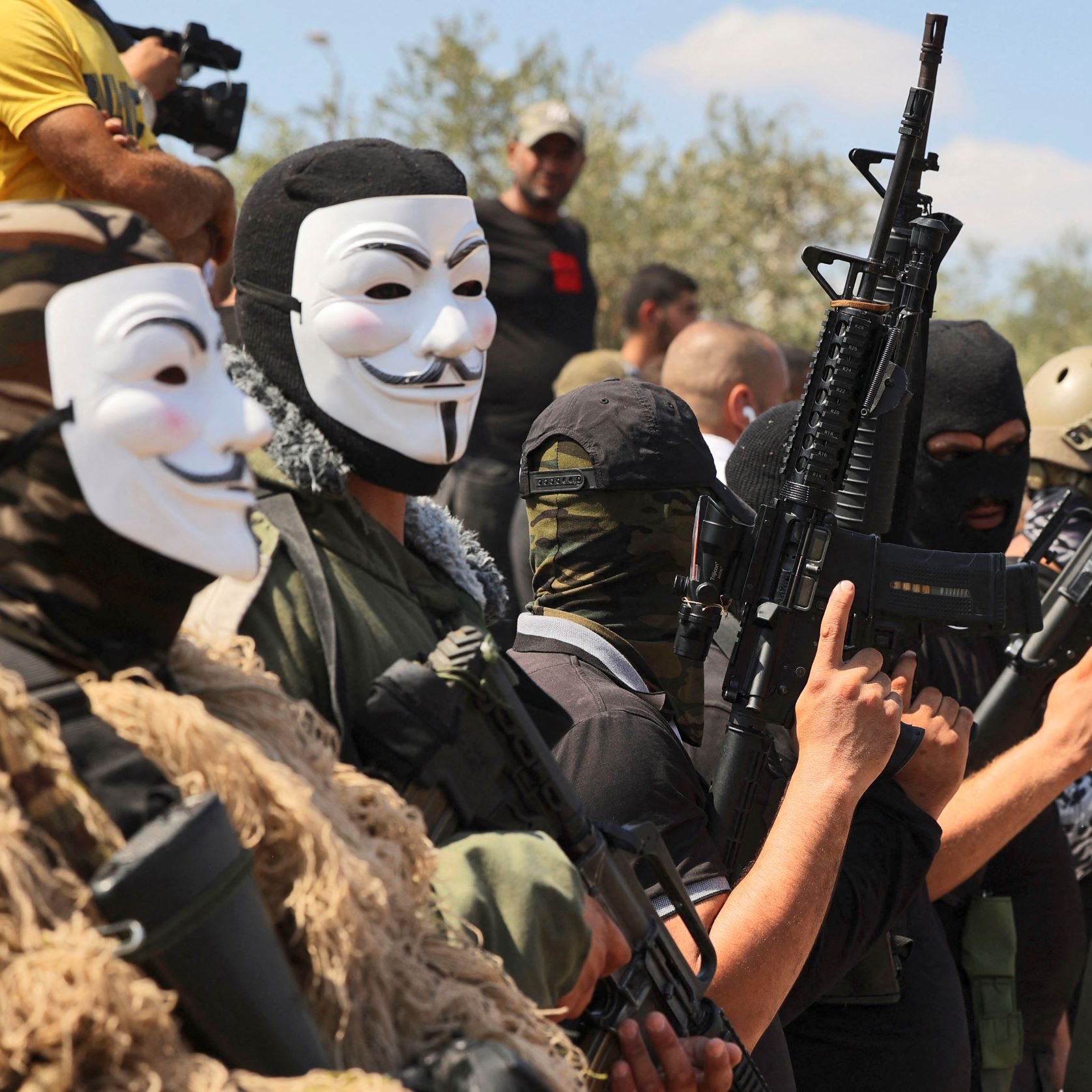 تظهر في الصورة مجموعة من الرجال يرتدون أقنعة وأزياء عسكرية، ويحمون أسلحة متنوعة. خلفهم يوجد عدد من الأشخاص في الخلفية. تعكس الصورة أجواء عسكرية أو استعراض لمجموعة مسلحة، حيث يتم التركيز على أسلحتهم وتنسيقهم. السماء تبدو زرقاء مع بعض السحاب، مما يعطي انطباعًا عن الأجواء المحيطة.