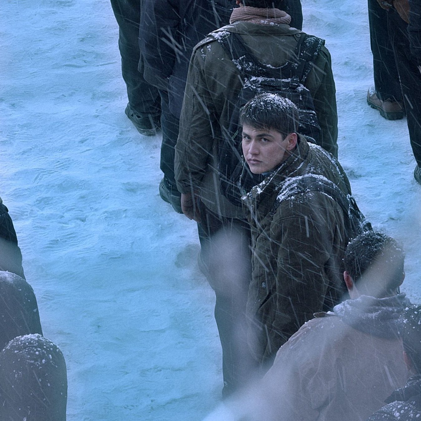 L'image montre une scène froide et enneigée avec une foule de personnes, des flocons de neige tombant. Au centre, un jeune homme se retourne, son visage est légèrement visible. Les autres autour de lui sont flous et vêtus de manteaux sombres, créant une atmosphère de tension et d'incertitude.