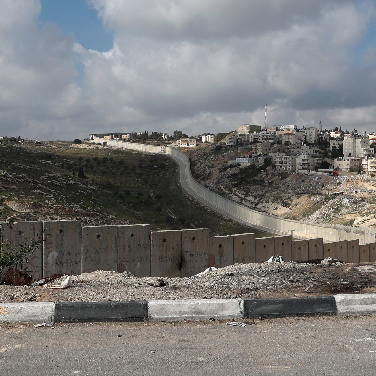 در این تصویر، یک پسر در حال پیاده‌روی در کنار دیواری بلند و سیم‌خاردار به نظر می‌رسد. این دیوار در زمینه‌ی یک منطقه شهری قرار دارد که ساختمان‌های مسکونی در آن دیده می‌شود. آسمان ابری است و ابرها در حال حرکت هستند. فضای تصویر ممکن است احساساتی از تنهایی و جدایی را منتقل کند. جاده‌ای در جلوی تصویر وجود دارد و محیط به‌نظر خشک و ناآرام می‌آید.