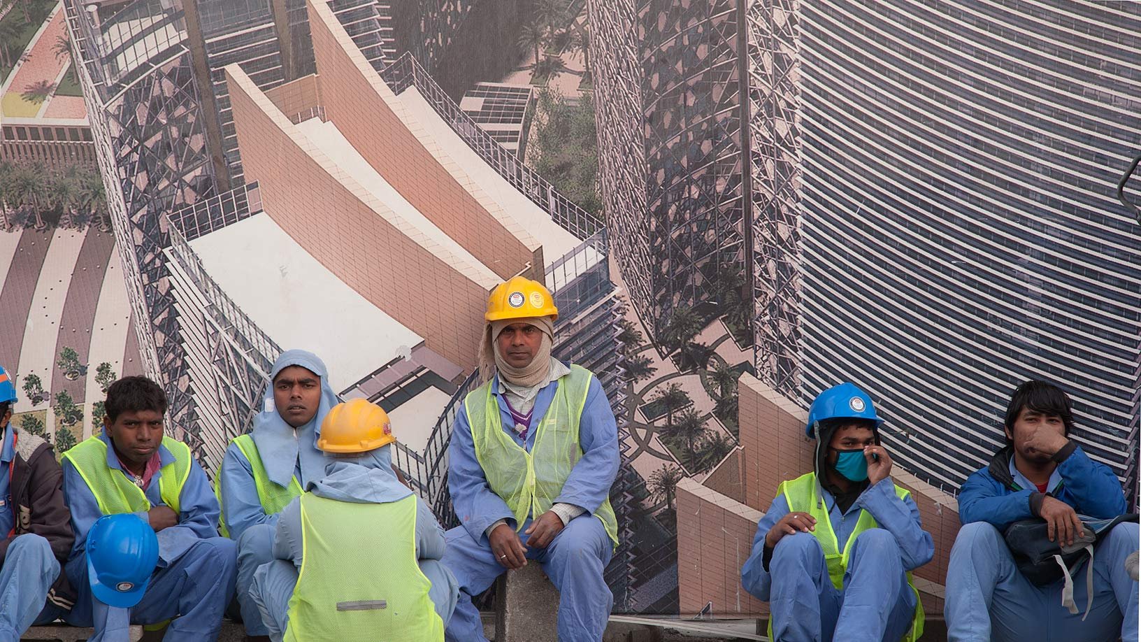 En la imagen se pueden ver a varios trabajadores de la construcción sentados sobre una superficie. Están vistiendo uniformes azules y chalecos reflectantes. Algunos llevan casco de diferentes colores, como amarillo y azul. Al fondo, hay un gran cartel que muestra una imagen de un edificio moderno, sugiriendo que están trabajando en un proyecto de construcción. Los trabajadores parecen estar en un descanso, y el ambiente refleja una jornada laboral intensa.