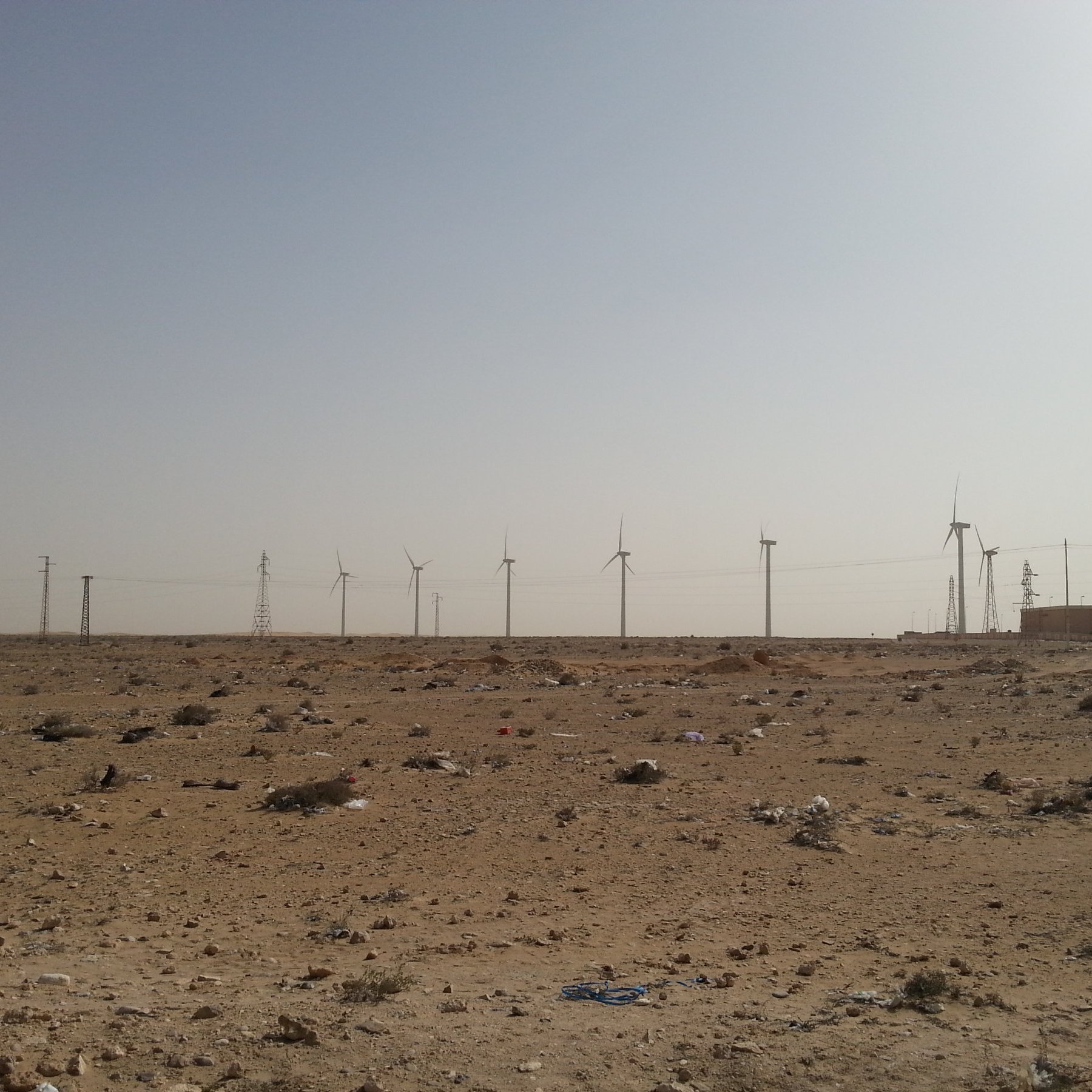 L'image montre un paysage désertique, caractérisé par un terrain aride avec des débris, probablement des déchets, éparpillés sur le sol. À l'arrière-plan, on peut voir plusieurs éoliennes qui s'élèvent vers le ciel clair. Des pylônes électriques sont également visibles, ajoutant une touche industrielle à ce paysage naturel. Le ciel est dégagé, ce qui accentue l'effet désertique de la scène.