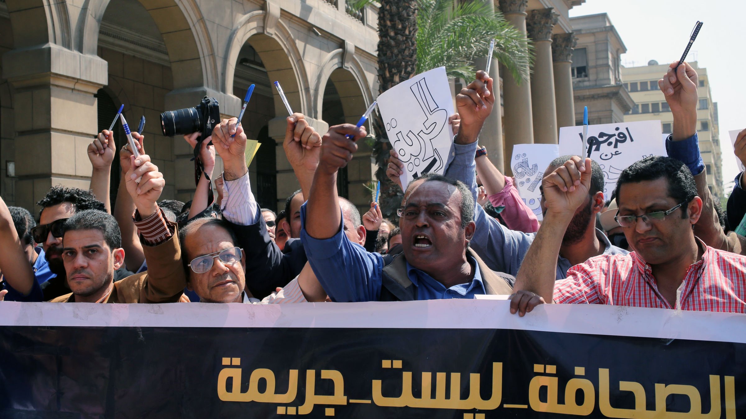 L'image montre une manifestation avec un groupe de personnes levant les bras et tenant des pancartes. Ils semblent exprimer leur mécontentement ou revendiquer des droits, notamment en rapport avec la liberté de la presse. L'atmosphère est énergique et déterminée, avec des visages montrant des émotions de passion et de revendication. À l'arrière-plan, on peut voir des bâtiments qui indiquent un environnement urbain. Le slogan sur la banderole semble défendre une position sur la presse, suggérant que celle-ci ne devrait pas être considérée comme un crime.