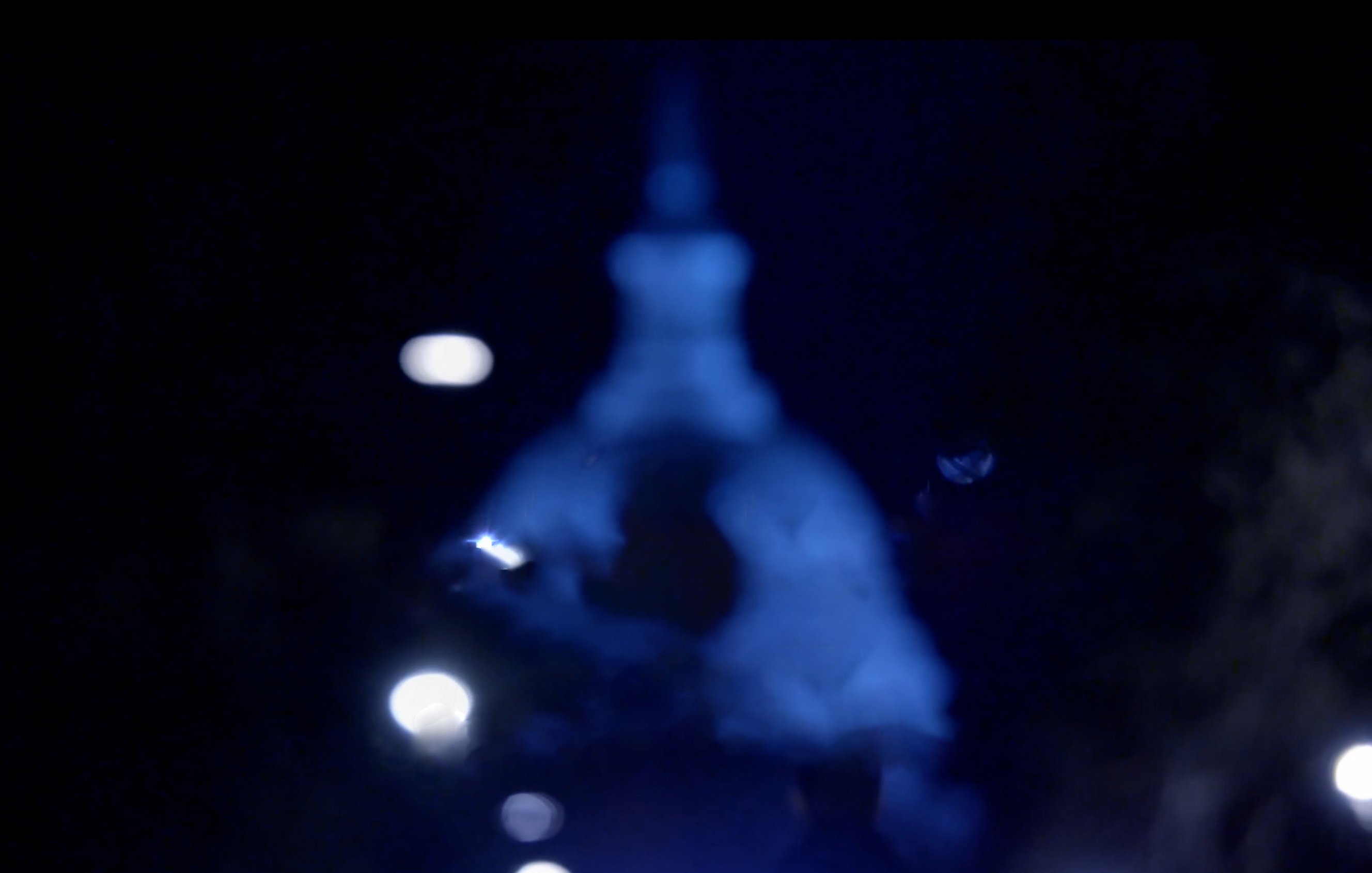 L'image montre une scène nocturne avec un éclairage diffus. Au centre, on aperçoit une silhouette floue, probablement d'un bâtiment ou d'une structure, avec un dôme et une pointe. Les lumières autour ajoutent une ambiance mystérieuse et éthérée, créant un contraste entre l'obscurité environnante et les sources lumineuses. L'atmosphère semble calme et légèrement mélancolique.