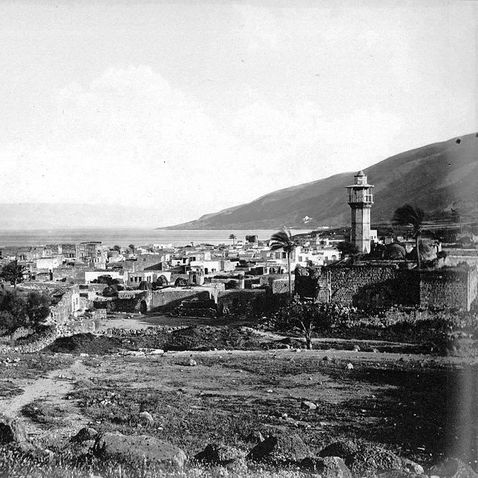 The image depicts a historical view of a coastal town near a body of water, likely a lake or sea, with a prominent lighthouse tower standing tall in the background. The landscape features a mix of stone buildings and ruins, suggesting a blend of architecture that may reflect the area's history. Surrounding the town are hills or mountains, adding depth to the scene. The overall mood is serene and nostalgic, capturing a moment from the past with a monochromatic or sepia tone.
