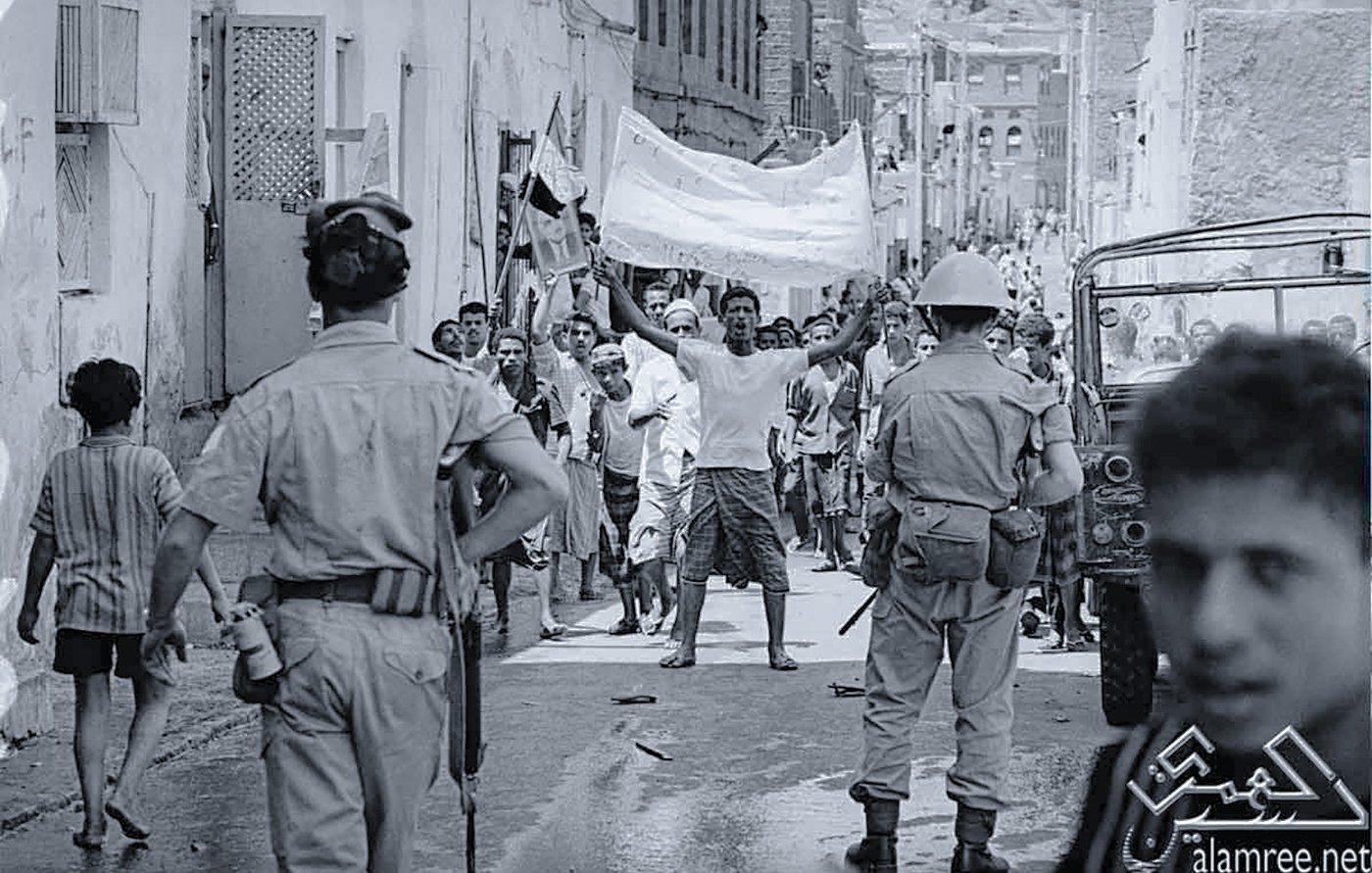 L'image montre une scène de rue dans un contexte de manifestation ou de rassemblement. On peut voir des groupes de personnes, certains brandissant des drapeaux ou des pancartes, exprimant visiblement leur émotion ou leur revendication. Des policiers réglementent la situation, et l'atmosphère semble tendue. L'environnement urbain est composé de bâtiments simples, typiques d'une ville. Les vêtements des gens indiquent peut-être une époque passée, soulignant le contexte historique de la scène.
