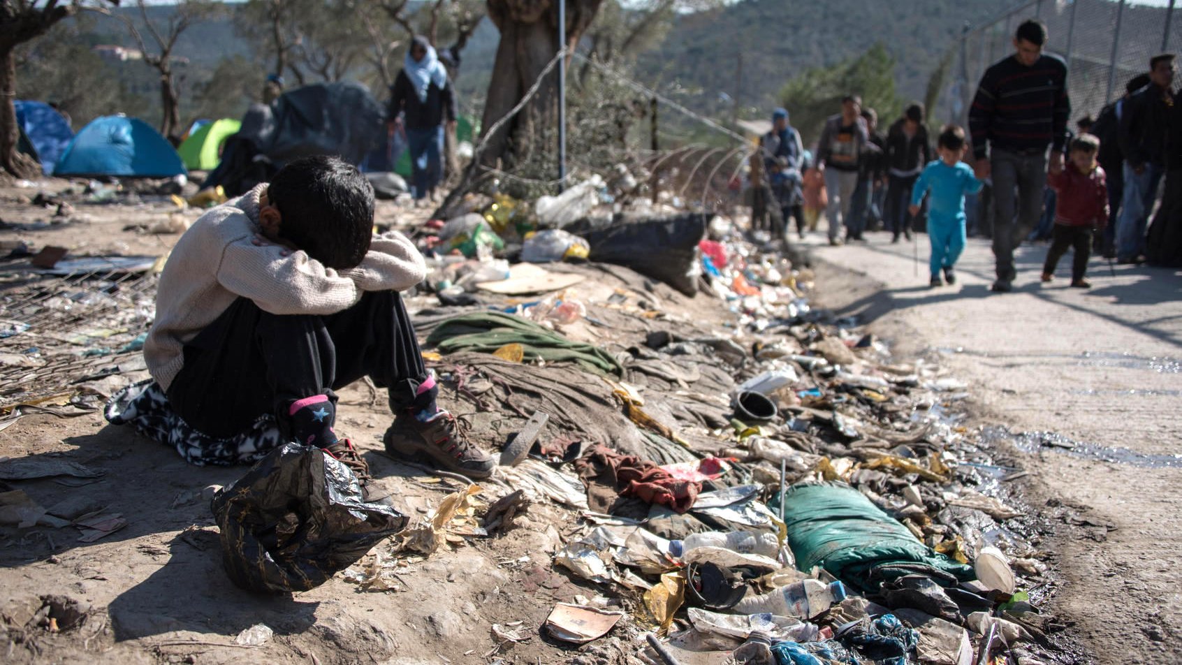 L'image montre un jeune enfant assis au sol, semblant triste ou désemparé. Il est entouré de déchets et de débris, ce qui souligne une situation de pauvreté et de désespoir. À l'arrière-plan, on peut voir d'autres personnes marchant, probablement des familles, dans un environnement difficile. Des tentes sont visibles, indiquant un campement temporaire. L'ensemble de la scène évoque une réalité difficile et poignante.