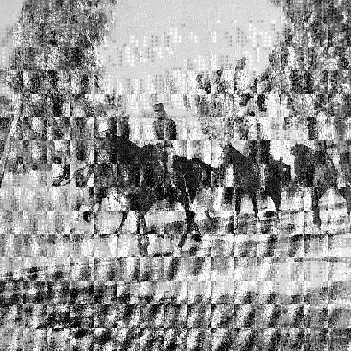 L'image montre un groupe de cavaliers, probablement des soldats, sur des chevaux, défilant sur une route. Il y a des arbres sur les côtés, et au premier plan, on peut voir une personne marchant sur le côté du chemin. À l'arrière-plan, des bâtiments et un clocher sont visibles, suggérant un environnement urbain. L'atmosphère semble historique, peut-être photographiée au début du XXe siècle.