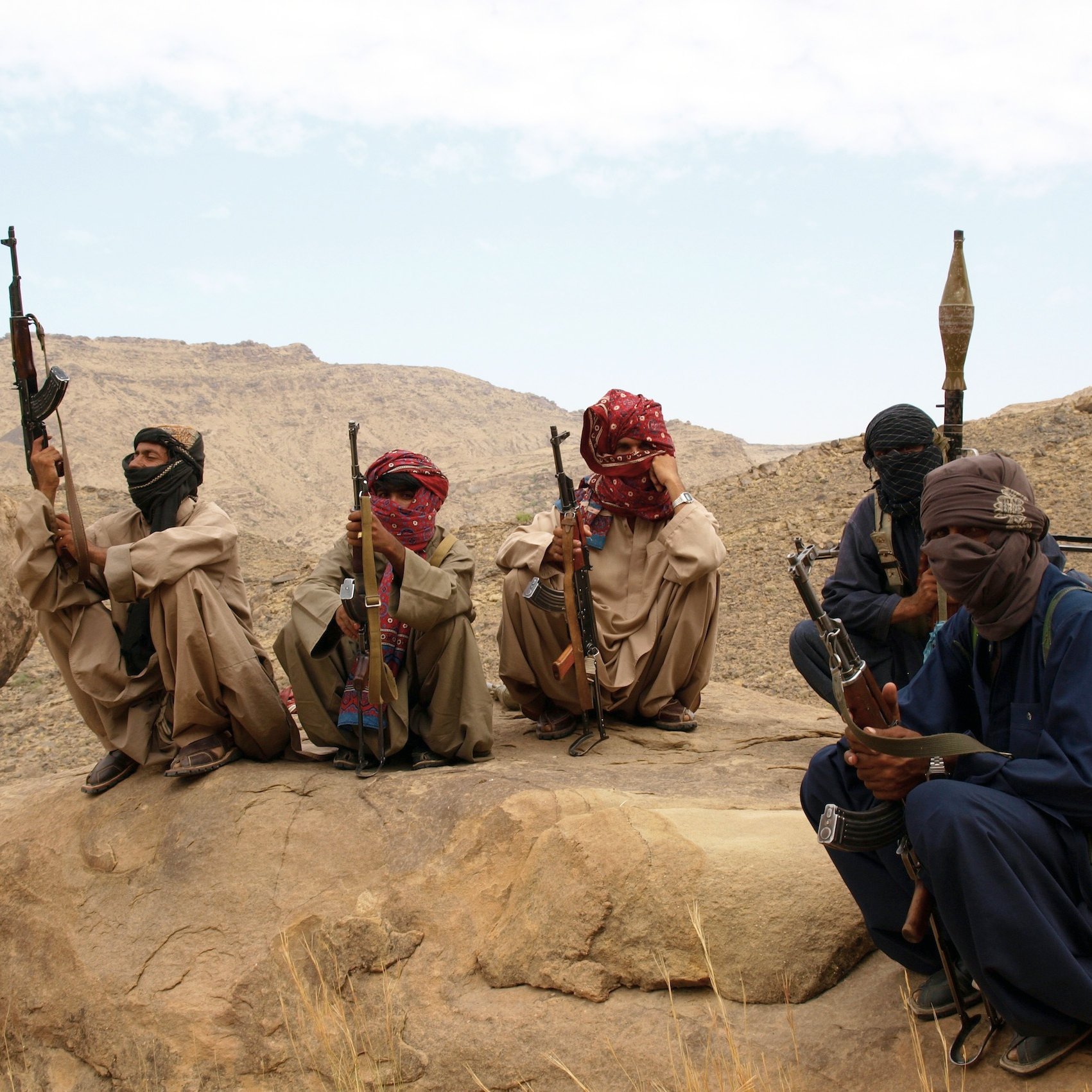 L'image montre un groupe de personnes assises sur des rochers dans un paysage désertique. Elles portent des vêtements traditionnels, masquent partiellement leur visage avec des foulards et tiennent des armes. L'environnement est montagneux et aride, avec des formations rocheuses en arrière-plan. L'ambiance semble sérieuse et reflète une situation de tension.