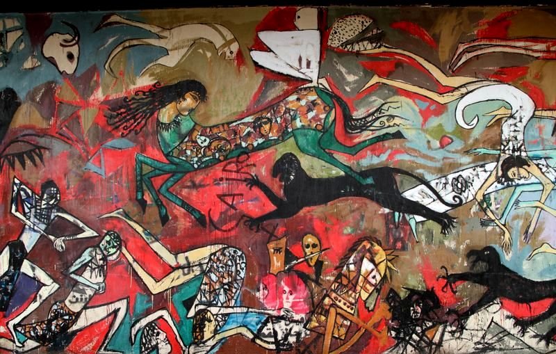 L'image représente une fresque murale vibrante et dynamique, riche en couleurs et en motifs. On y voit des figures humaines stylisées, certaines en mouvement, qui semblent interagir dans un environnement chaotique. Les silhouettes sont variées, avec des éléments abstraits et des formes aux contours fluides. Des éléments symboliques tels que des animaux et des motifs géométriques ajoutent à la complexité de l'œuvre. L'ensemble dégage une atmosphère à la fois énergique et mystique, témoignant d'une forte expression artistique.