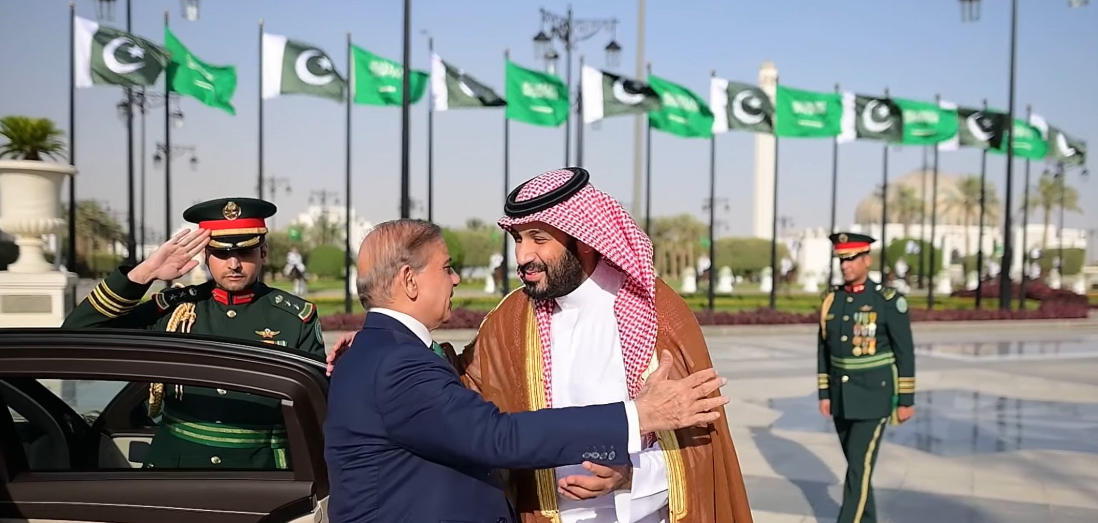A formal greeting between two leaders, with guards and flags in the background.