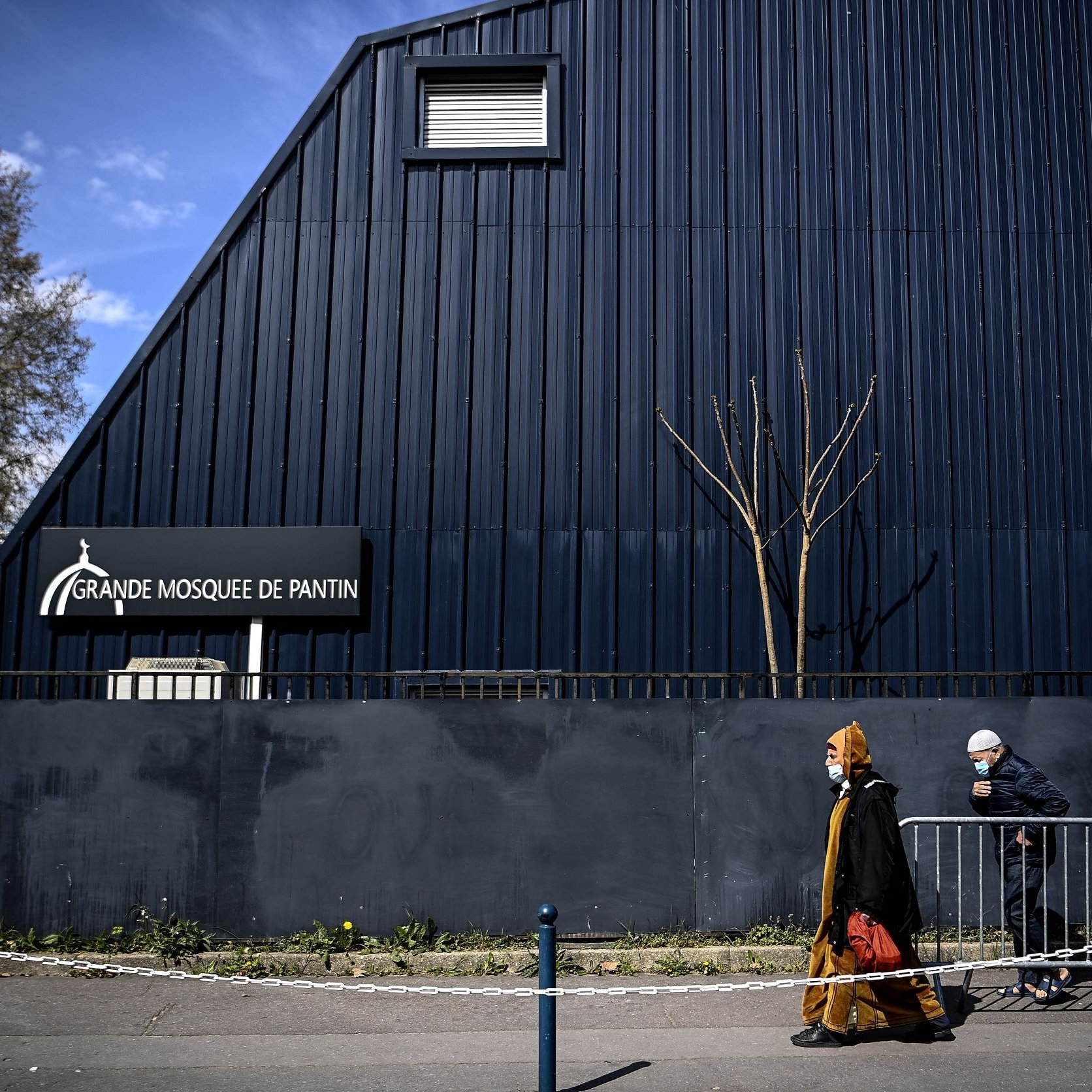 L'image montre une scène extérieure près d'un bâtiment. On peut voir la façade d'une grande mosquée, identifiée par un panneau indiquant "Grande Mosquée de Pantin". À l'avant-plan, deux personnes marchent le long d'un chemin, l'une d'elles portant un voile et une tenue traditionnelle, tandis que l'autre est vêtue d'une manière plus décontractée, portant également un masque. L'architecture du bâtiment est moderne, avec un toit incliné, et l'environnement semble urbain, avec quelques arbres dépouillés de feuilles. Une barrière de sécurité sépare la rue du bâtiment.