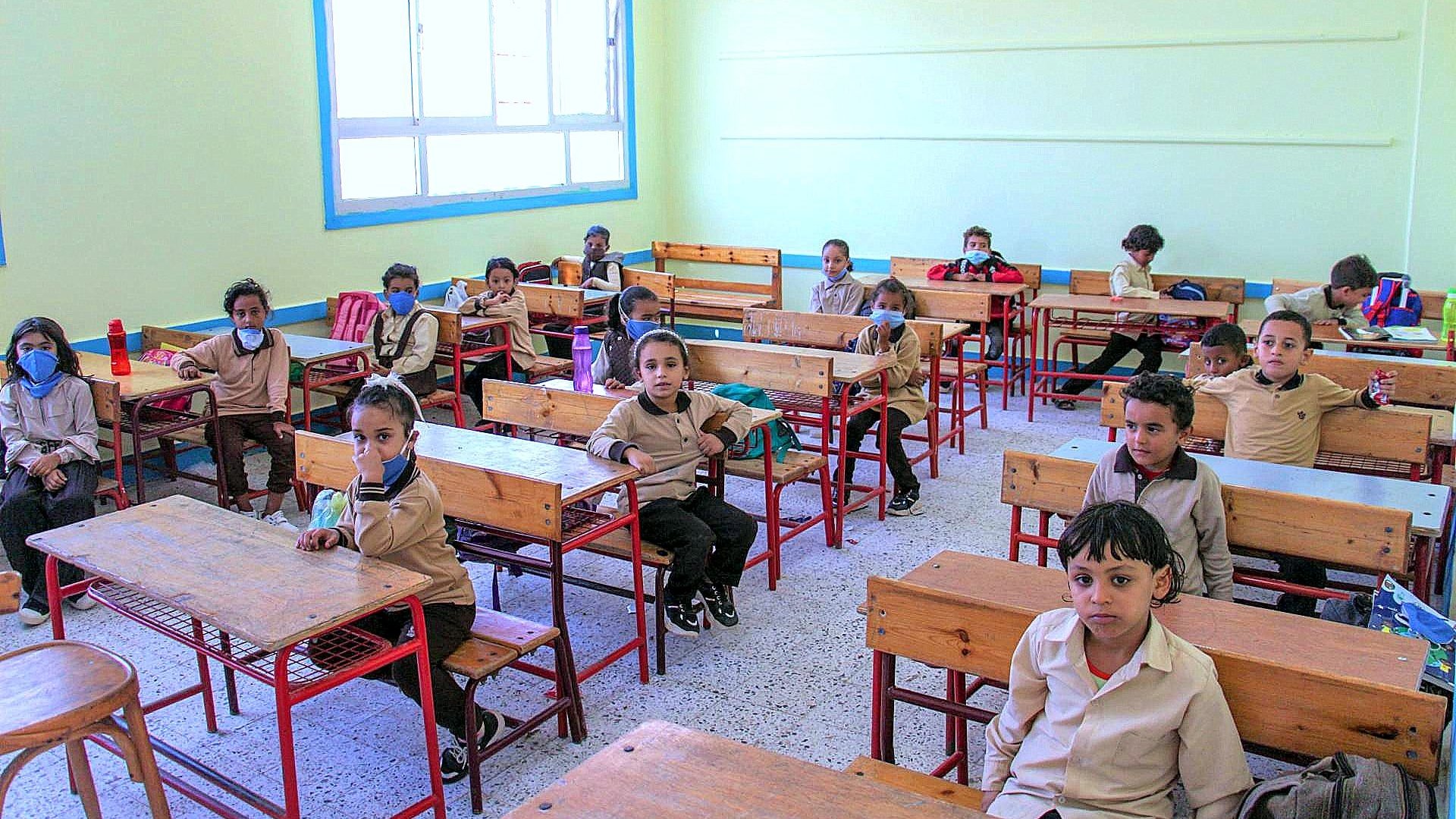 L'image montre une salle de classe avec plusieurs élèves assis à des bureaux en bois. Les enfants portent des uniformes beige et plusieurs d'entre eux ont des masques sur le visage. La pièce est lumineuse, avec des murs peints en jaune clair et des fenêtres qui laissent entrer la lumière. Les élèves semblent attentifs, certains regardant vers l'appareil photo. Les bureaux sont espacés, et il y a quelques fournitures scolaires visibles sur les tables.