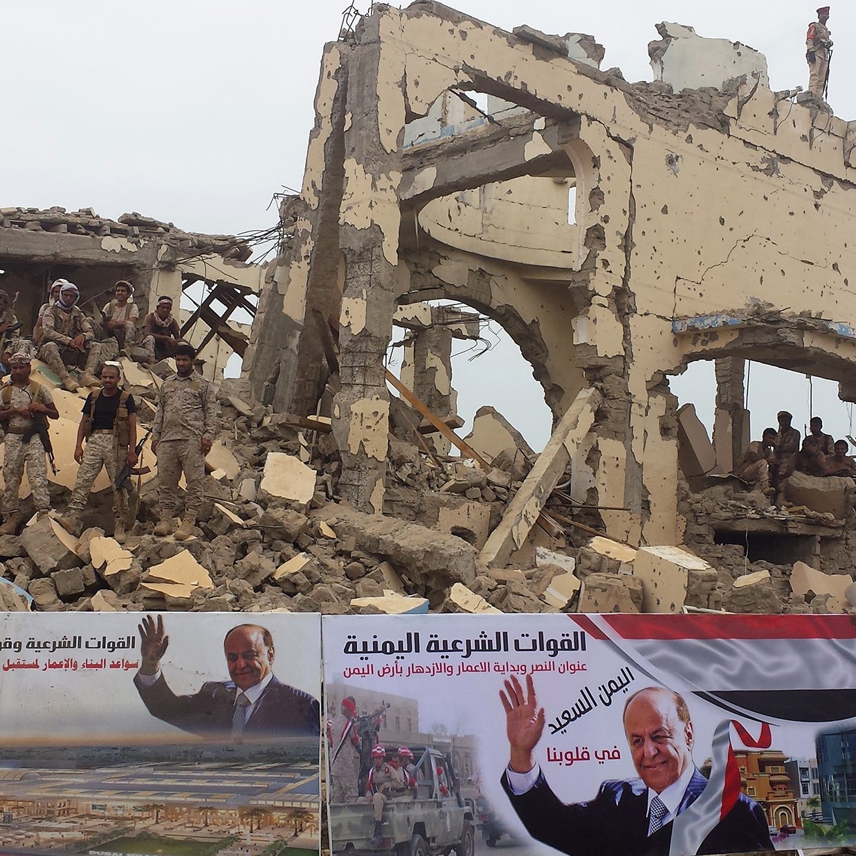 L'image montre un bâtiment en ruines, probablement à la suite d'un conflit ou d'une destruction. Des soldats sont visibles parmi les débris, ce qui indique une présence militaire dans la zone. Au premier plan, des affiches sont accrochées, probablement en soutien à un leader ou un mouvement, avec du texte en arabe. L'ensemble évoque une atmosphère de résilience et de reconstruction au milieu des dégâts.
