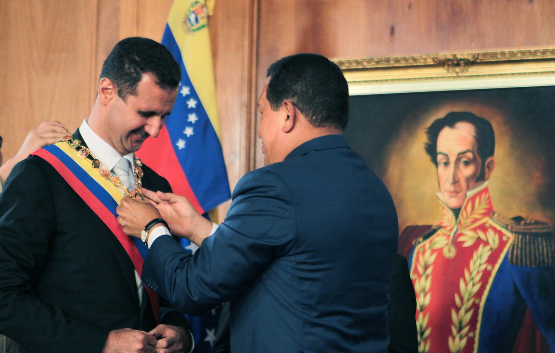 L'image montre une cérémonie officielle où un homme, probablement un leader politique, reçoit un insigne ou un collier de la part d'un autre homme. Le premier homme est vêtu d'un costume noir et porte une écharpe, tandis que le second, habillé en bleu, est en train de lui remettre l'insigne autour du cou. En arrière-plan, on aperçoit un portrait d'une figure historique, ainsi que des drapeaux, ce qui suggère un contexte politique et national.