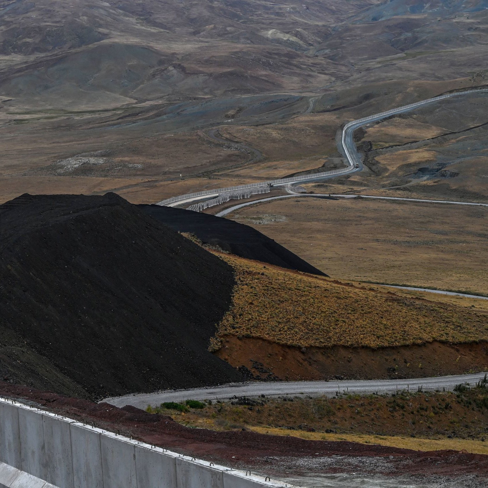 این تصویر نمایی از یک جاده در میان مناظر طبیعی خشک و کوهستانی را نشان می‌دهد. جاده به صورت منحنی و پیچ‌درپیچ از روی زمین‌های ناهموار عبور می‌کند و در سمت راست تصویر، دیواری سفیدرنگ وجود دارد که به طور راستا در حال پیشروی است. در پس‌زمینه، تپه‌ها و دامنه‌های کوهستانی با رنگ‌های ملایم و خاصی دیده می‌شوند که نمایی طبیعی و بکر را به تصویر می‌بخشند. فضای کلی تصویر به نوعی حس دورافتادگی و خلوتی را منتقل می‌کند.