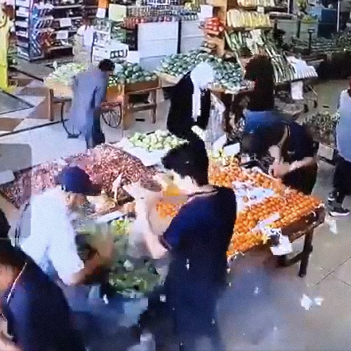 L'immagine mostra l'interno di un negozio di alimentari, probabilmente una fruttivendolo. Ci sono diverse persone che si muovono tra le corsie, mentre vari tipi di frutta e verdura sono esposti su tavoli e scaffali. Le persone sembrano occupate a scegliere e acquistare prodotti freschi. La scena è vivace, con molti colori provenienti dai frutti esposti, come agrumi e verdure di vario tipo. La disposizione degli oggetti suggerisce un'ambientazione ordinata e ben fornita.