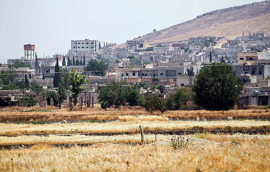 تظهر الصورة منطقة ريفية حيث يتواجد عدد من المنازل والمباني على تلة. في الخلفية، يمكن رؤية جبال منخفضة، بينما في المقدمة يوجد حقل جاف بألوان ذهبية. تبرز بعض الأشجار والنباتات هنا وهناك، مما يضفي لمسة من الخضرة على المشهد. كما يوجد خزان مياه في الجانب الأيمن من الصورة، مما يشير إلى سكان المنطقة. الجو يبدو مشمسًا مع أجواء صيفية.
