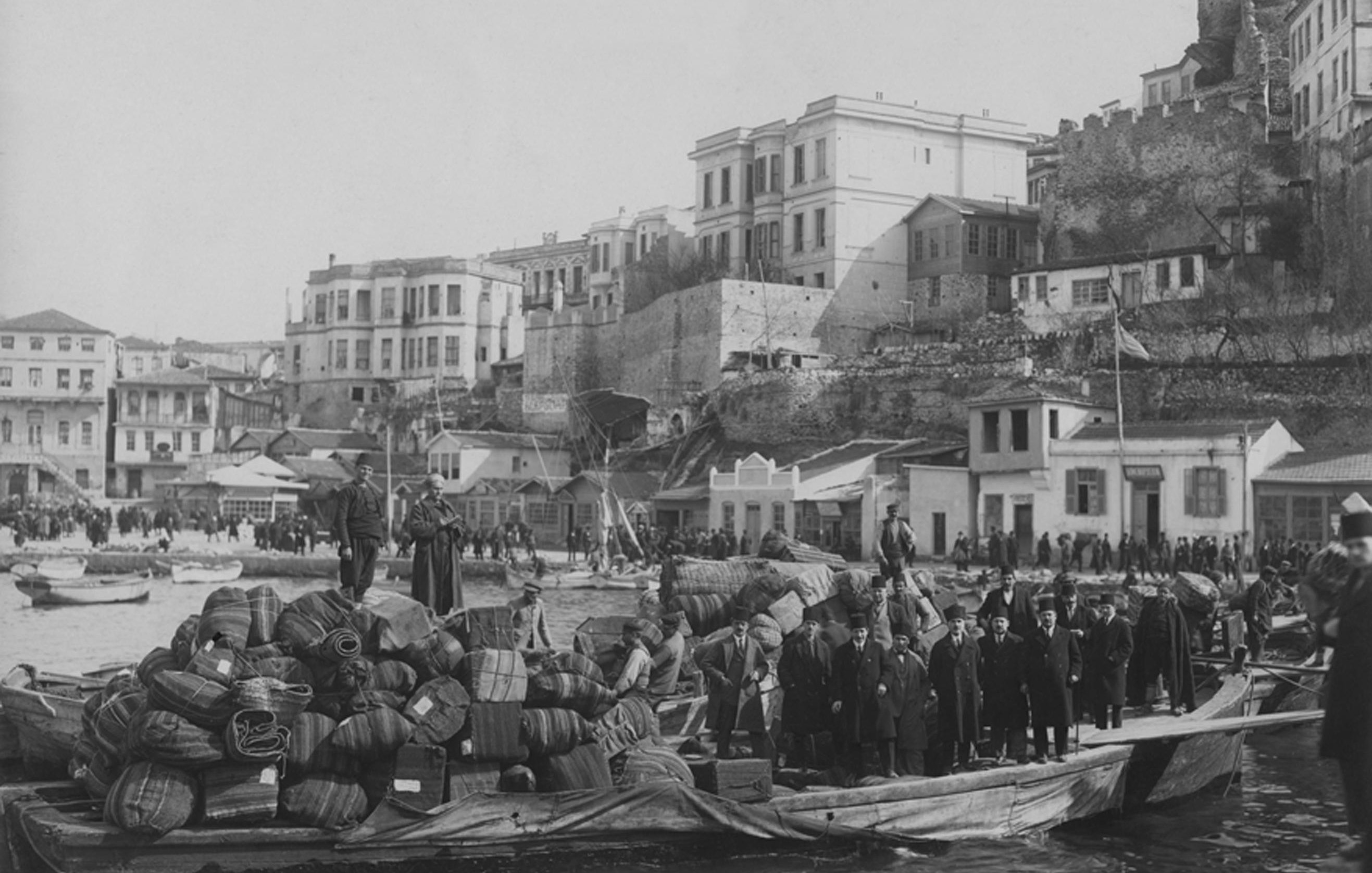 La imagen muestra un puerto animado en un contexto histórico, con un barco de madera cargado de bultos y barriles en primer plano. Un grupo de personas, posiblemente trabajadores o comerciantes, está de pie sobre la embarcación. Al fondo, se puede ver una serie de edificios y una muralla de piedra que se eleva, sugiriendo la presencia de una ciudad costera. La escena tiene un aspecto antiguo, lo que sugiere que fue tomada en una época pasada, posiblemente a finales del siglo XIX o principios del XX. Las actividades portuarias y el diseño arquitectónico transmiten una sensación de vida cotidiana en el puerto.