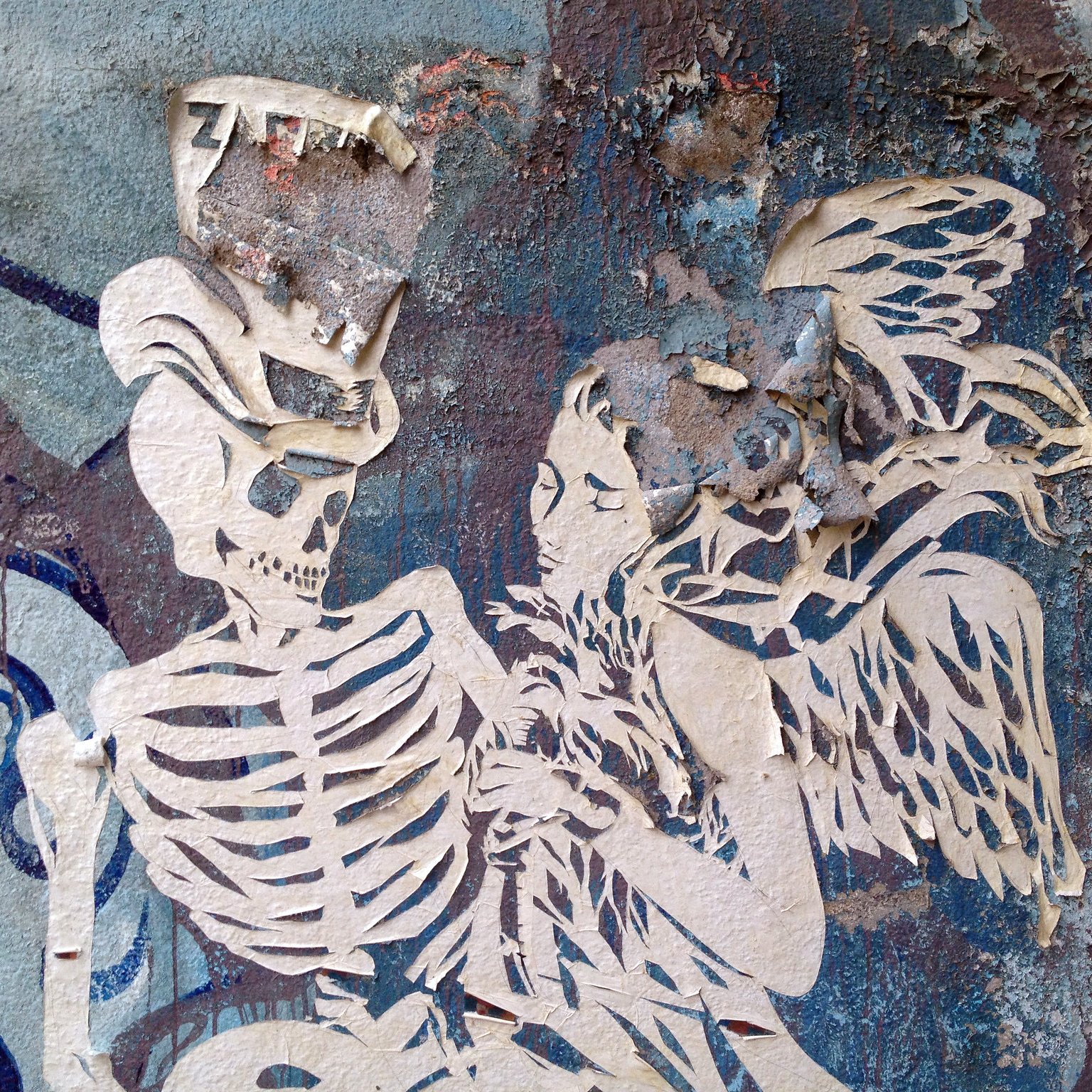 L'image montre une œuvre de street art sur un mur usé. On peut y voir un squelette et une figure féminine aux ailes, tous deux découpés dans un matériau qui semble être du papier ou du carton. Le fond est composé de textures et de couleurs dégradées, avec des traces d'usure et de peeling. L'ensemble dégage une atmosphère mystérieuse et à la fois poétique, mettant en avant les thèmes de la vie et de la mort.