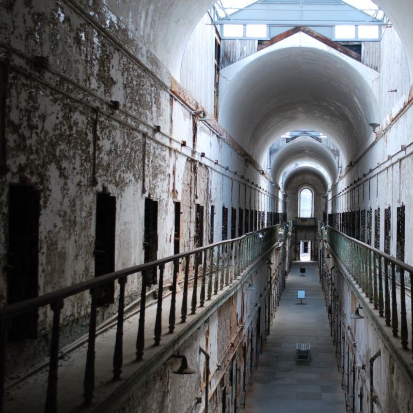 L'image montre l'intérieur d'une ancienne prison, avec un long couloir central bordé de cellules. Les murs sont décolorés et décrépis, témoignant de l'âge de l'établissement. Le plafond est haut, avec de grandes arches et une lumière qui pénètre par des lucarnes au-dessus. Les balustrades en fer forgé entourent les niveaux supérieurs, ajoutant à la structure vétuste. L'ambiance générale suggère une atmosphère d'abandon et d'histoire.